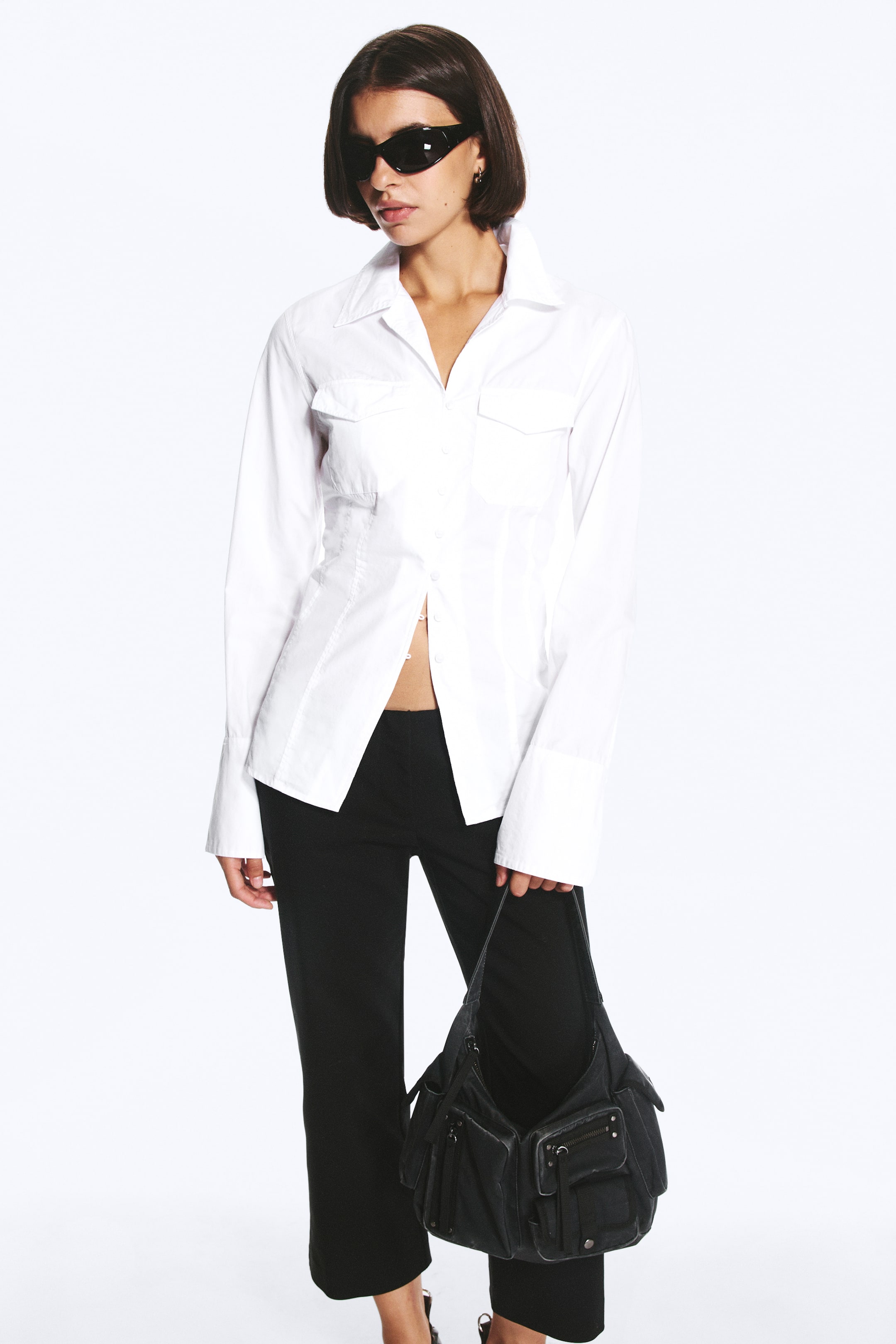 Προβολή μεγαλύτερης εικόνας: Ένα λευκό slim-fit πουκάμισο φοριέται από το μοντέλο, με μυτερό γιακά, τσέπες με καπάκι στο στήθος, ραφές διαμόρφωσης στη μέση και μακριά μανίκια με κλος μανσέτες. Το πουκάμισο είναι στυλαρισμένο μερικώς ξεκούμπωτο στη μέση με μαύρο παντελόνι, ενώ το μοντέλο κρατά μια μαύρη τσάντα χειρός.