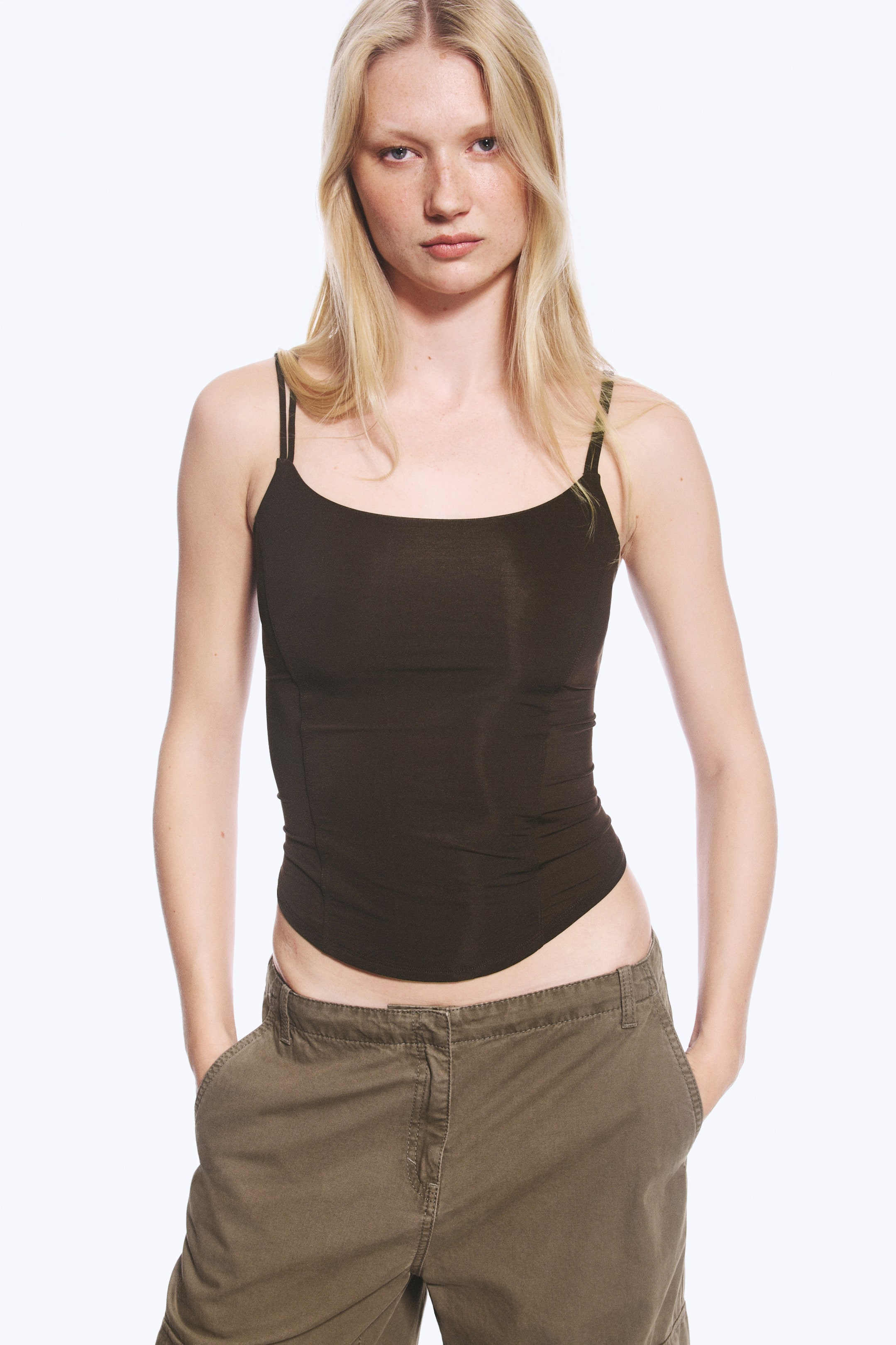 Se større billede: En mørkebrun corsetop med slim fit bæres af modellen og har en rund halsudskæring, tynde dobbelte skulderstropper og buede panelsømme langs kroppen. Toppen er designet med en buet kant og er stylet med olivengrønne bukser.