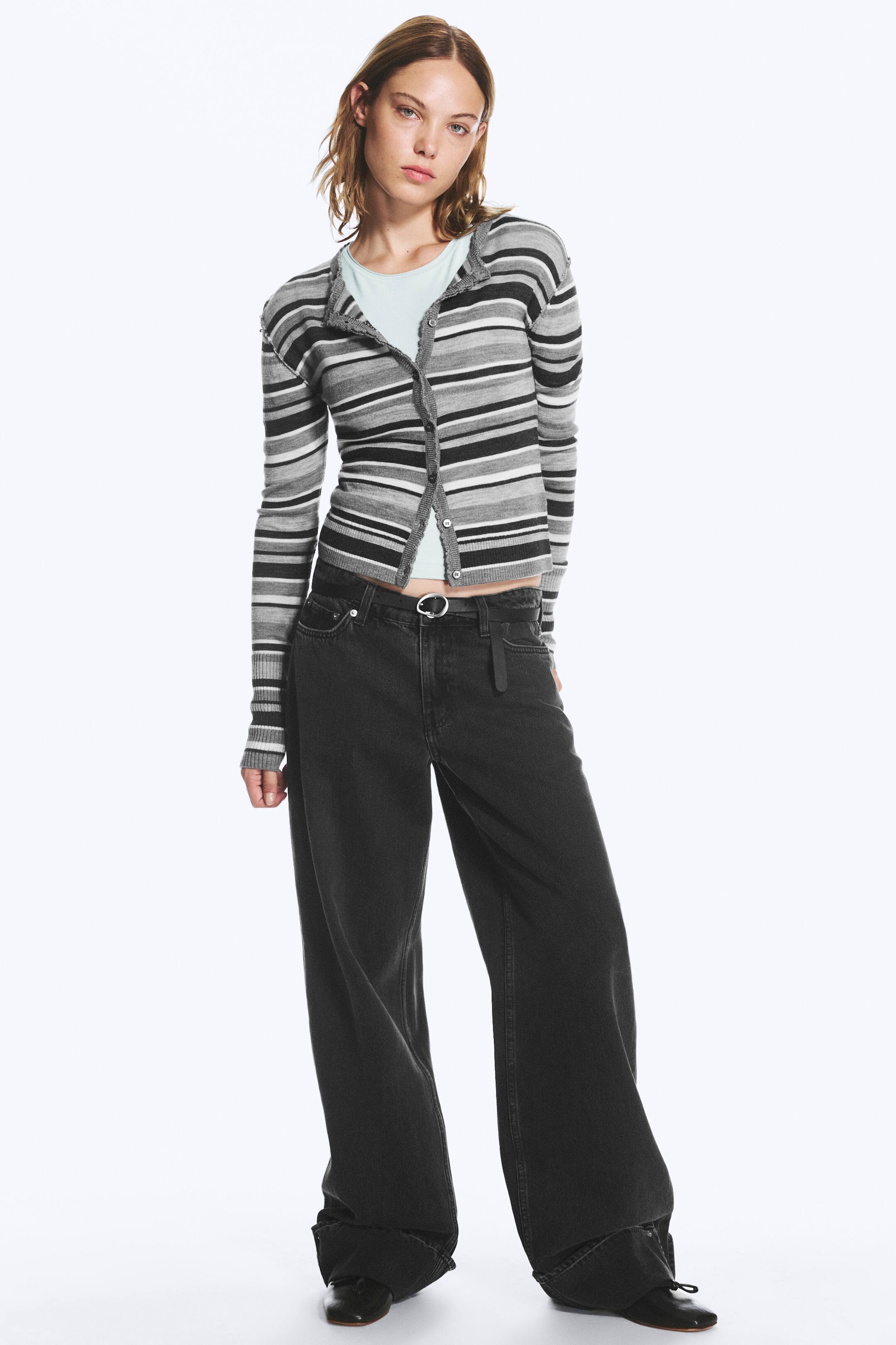 Grotere afbeelding bekijken: Een paar low-rise jeans met wijde pijpen in gewassen donkergrijs denim wordt gedragen door het model, met een losse pasvorm, klassieke five-pocket-styling en omgeslagen boorden aan de broekspijpen. De jeans zijn gestyled met een zwarte riem, een gestreept gebreid vest en zwarte schoenen.