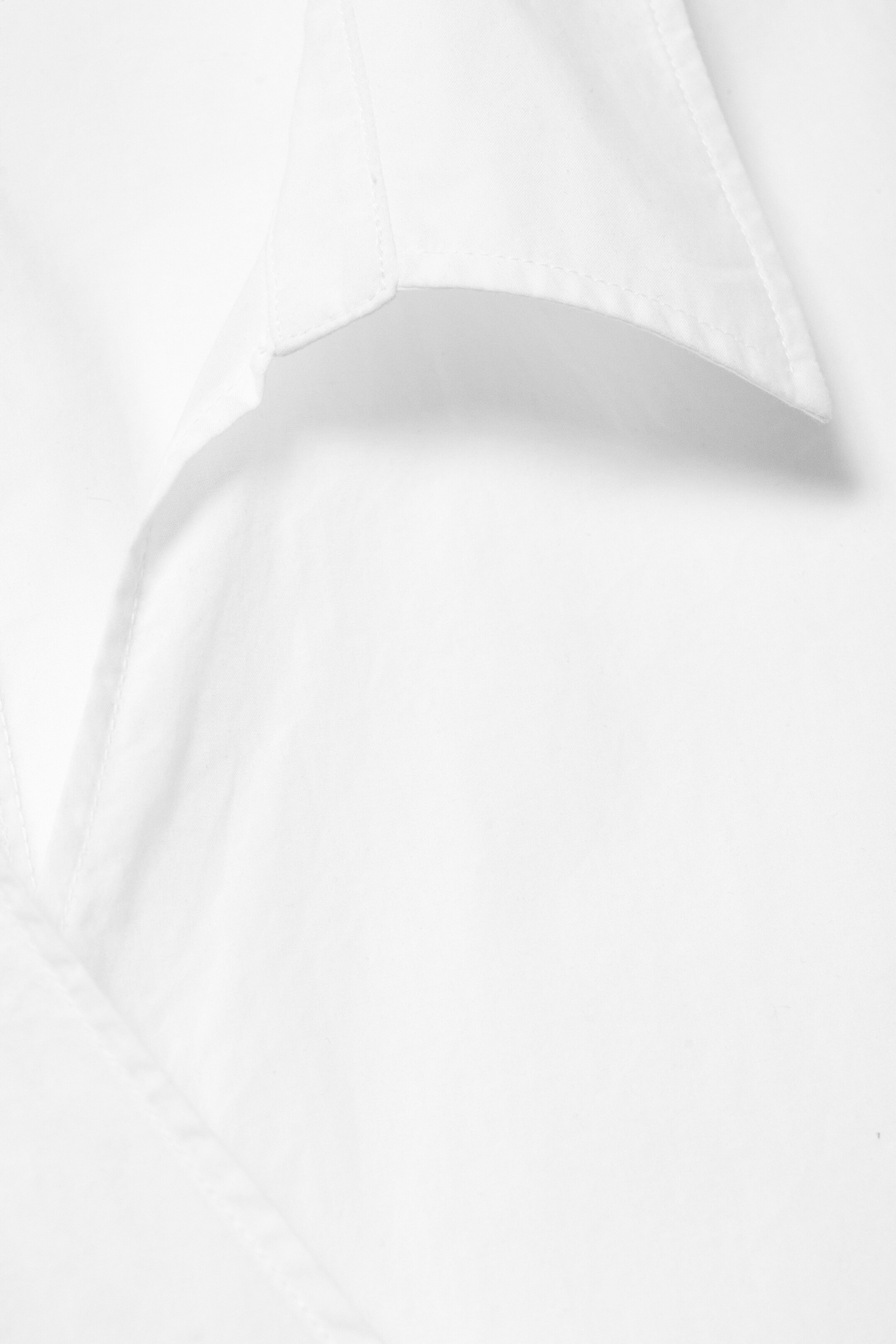 Agrandir l'image: Une chemise blanche présente un col à point ouvert avec des coutures visibles le long de ses bords. Le tissu de la chemise affiche des plis subtils et naturels.