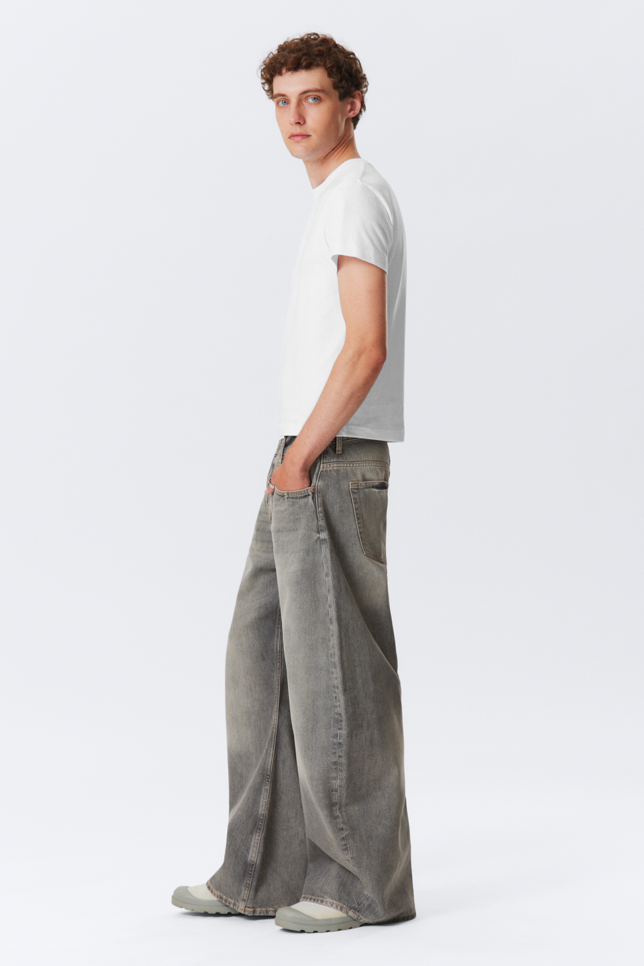 View larger image: A white short-sleeved t-shirt is being paired with loose-fitting, wide-leg grey jeans, presented from a side profile view. The jeans are featuring a faded wash and are extending fully to the ground, partially covering light-colored shoes.