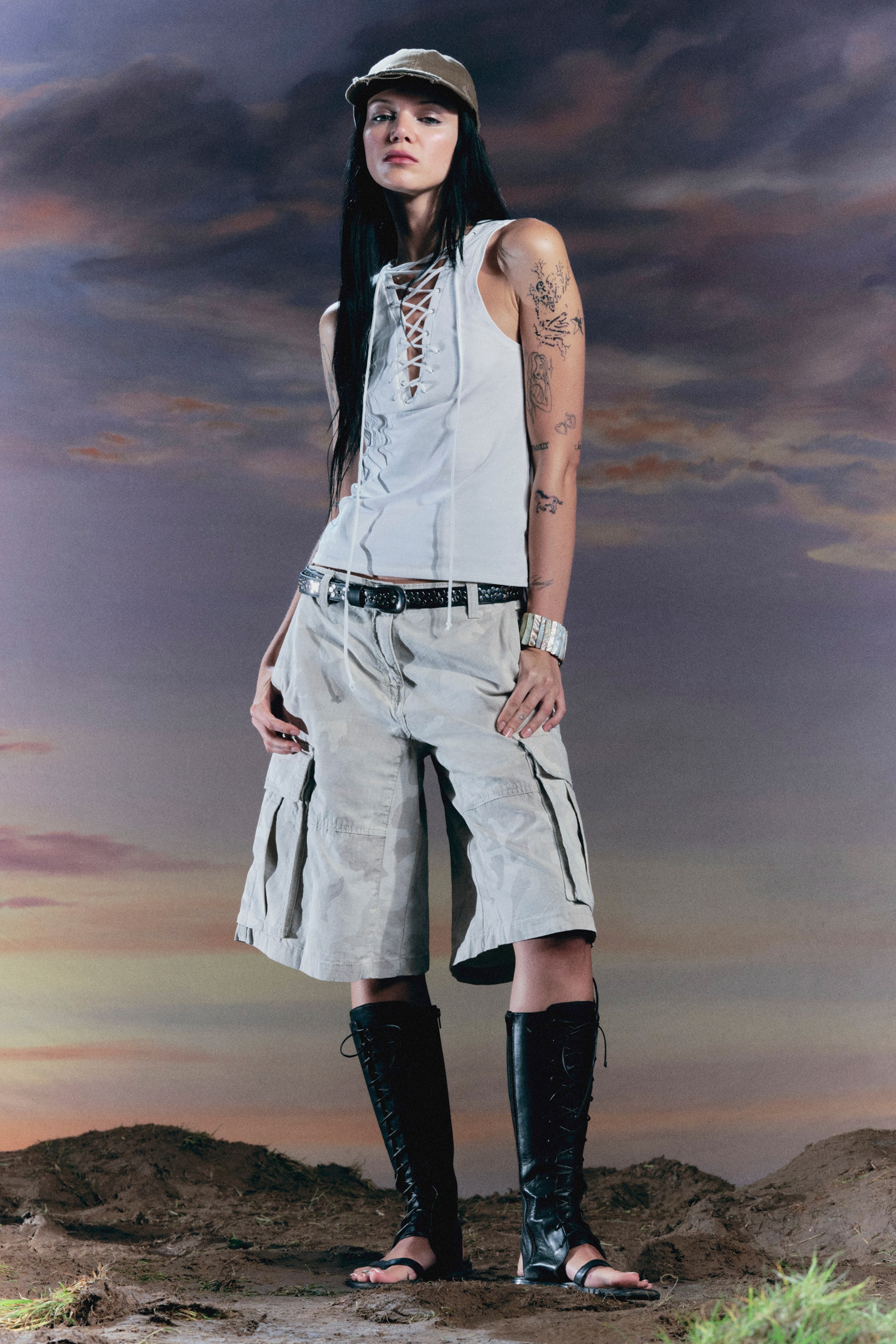 Näytä suurempi kuva: Vaaleansävyinen hihaton toppi, jossa on nyöritys edessä, yhdistettynä vaaleanbeigeihin maastokuvioituihin cargoshortseihin, joissa on sivutaskut ja musta koristeltu vyö. Hillitty oliivinvihreä lippalakki ja polvipituiset mustat nyörityksellä varustetut avokärkiset sandaalit täydentävät kokonaisuuden.