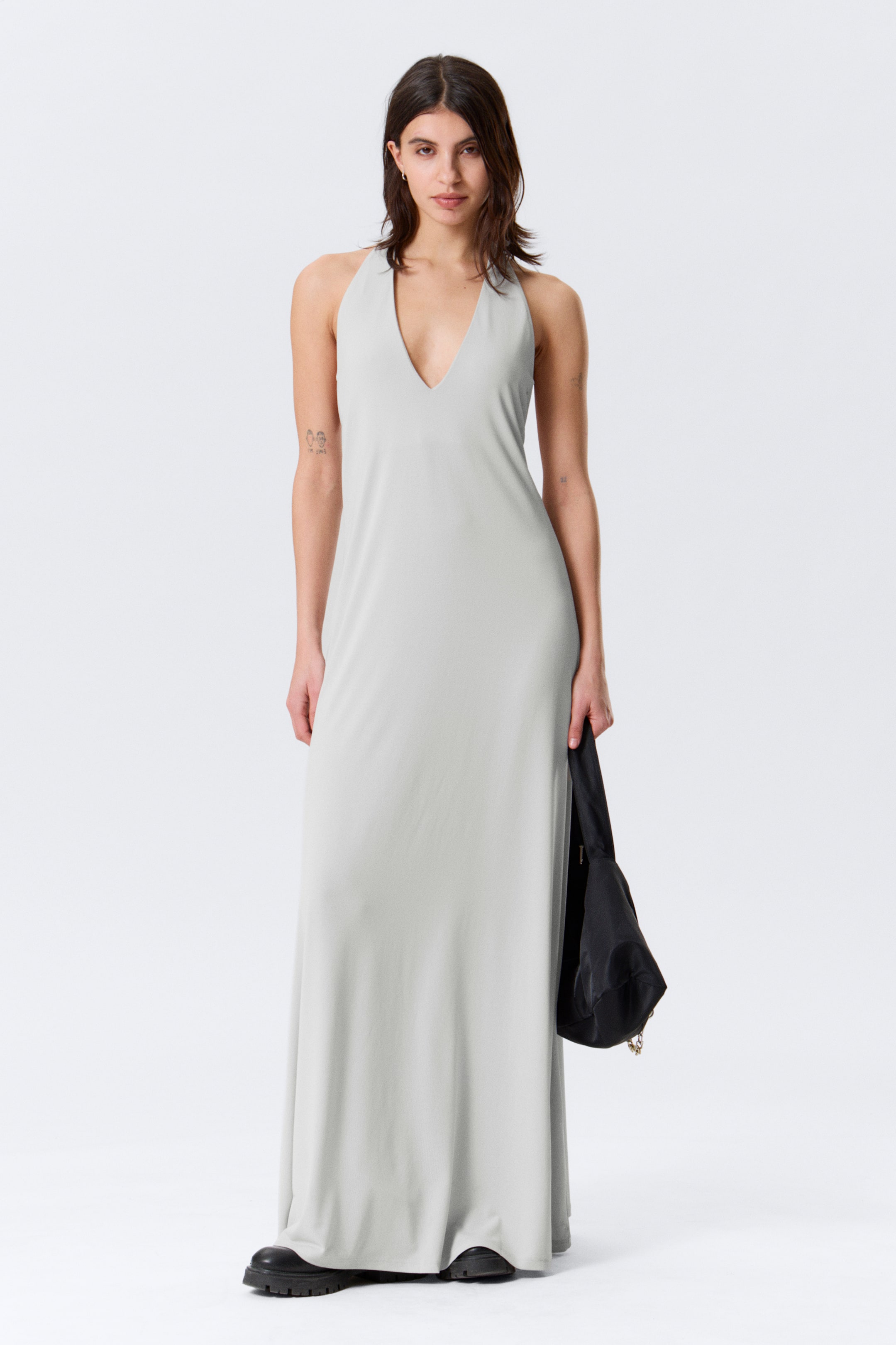 Se større billede: Den lysegrå maxikjole præsenterer en dyb V-udskæring og en halterneck, med stoffet der flyder glat ned til en gulvlængde kant.