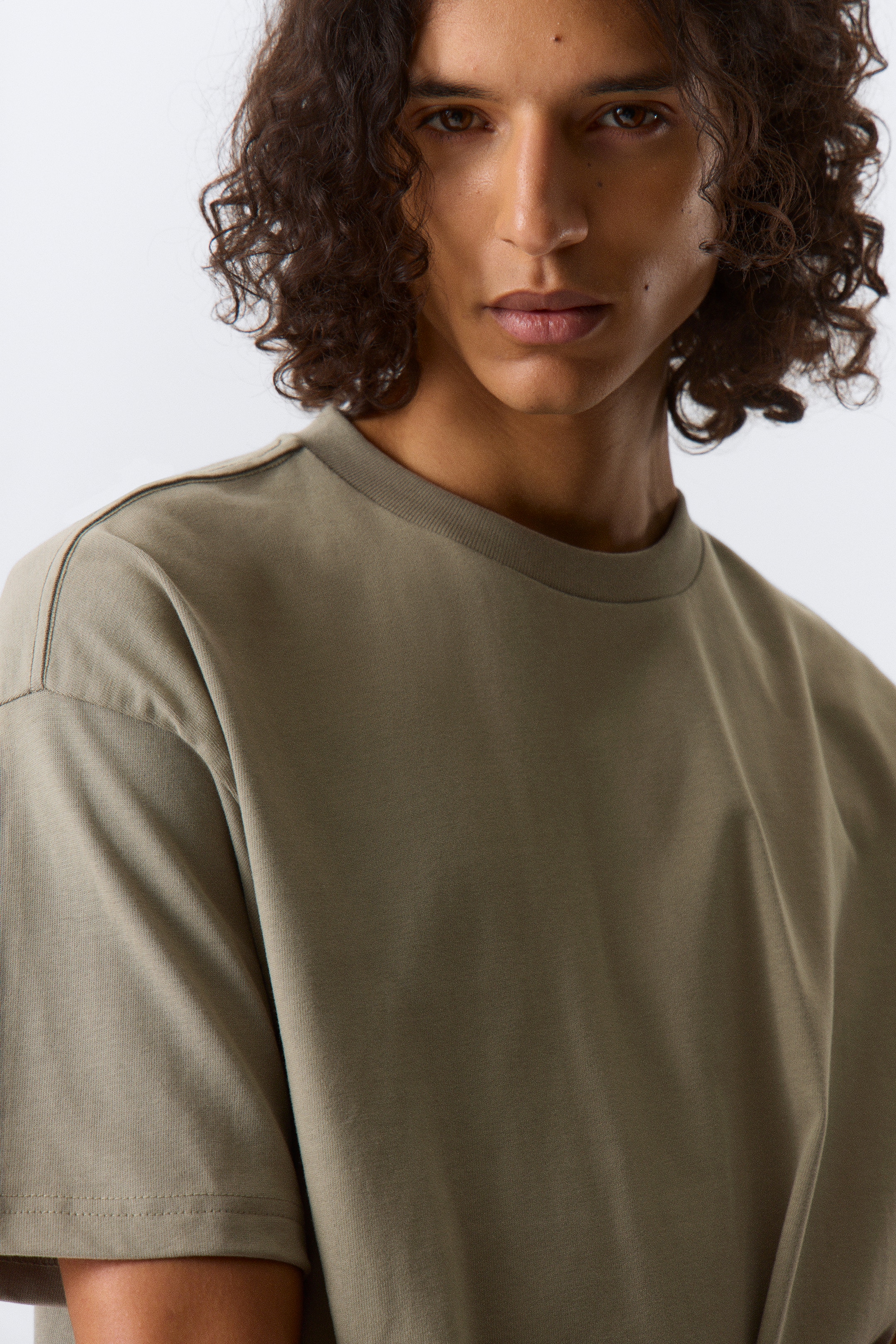 Grotere afbeelding bekijken: Een olijfgroen T-shirt met ronde hals wordt gedragen, enigszins schuin naar rechts. Het heeft korte, loszittende mouwen en zichtbare bovenstiksels langs de schoudernaden.