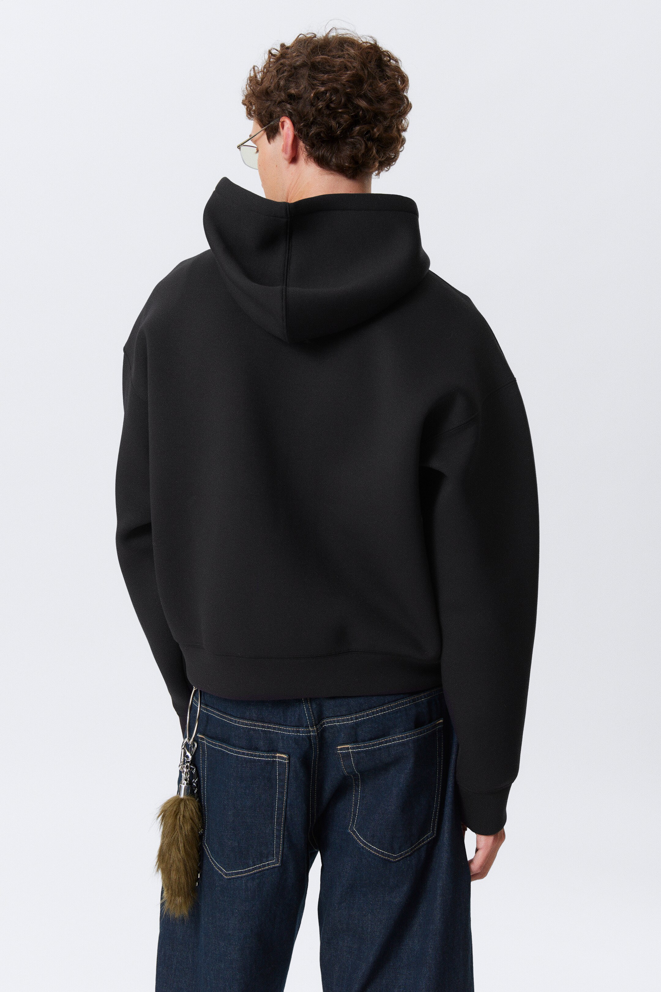 Ver imagem maior: Um hoodie preto está a ser mostrado de uma perspetiva traseira. Está a exibir um ajuste oversized e descontraído com ombros caídos, um capuz estruturado e uma faixa de bainha larga e plana.