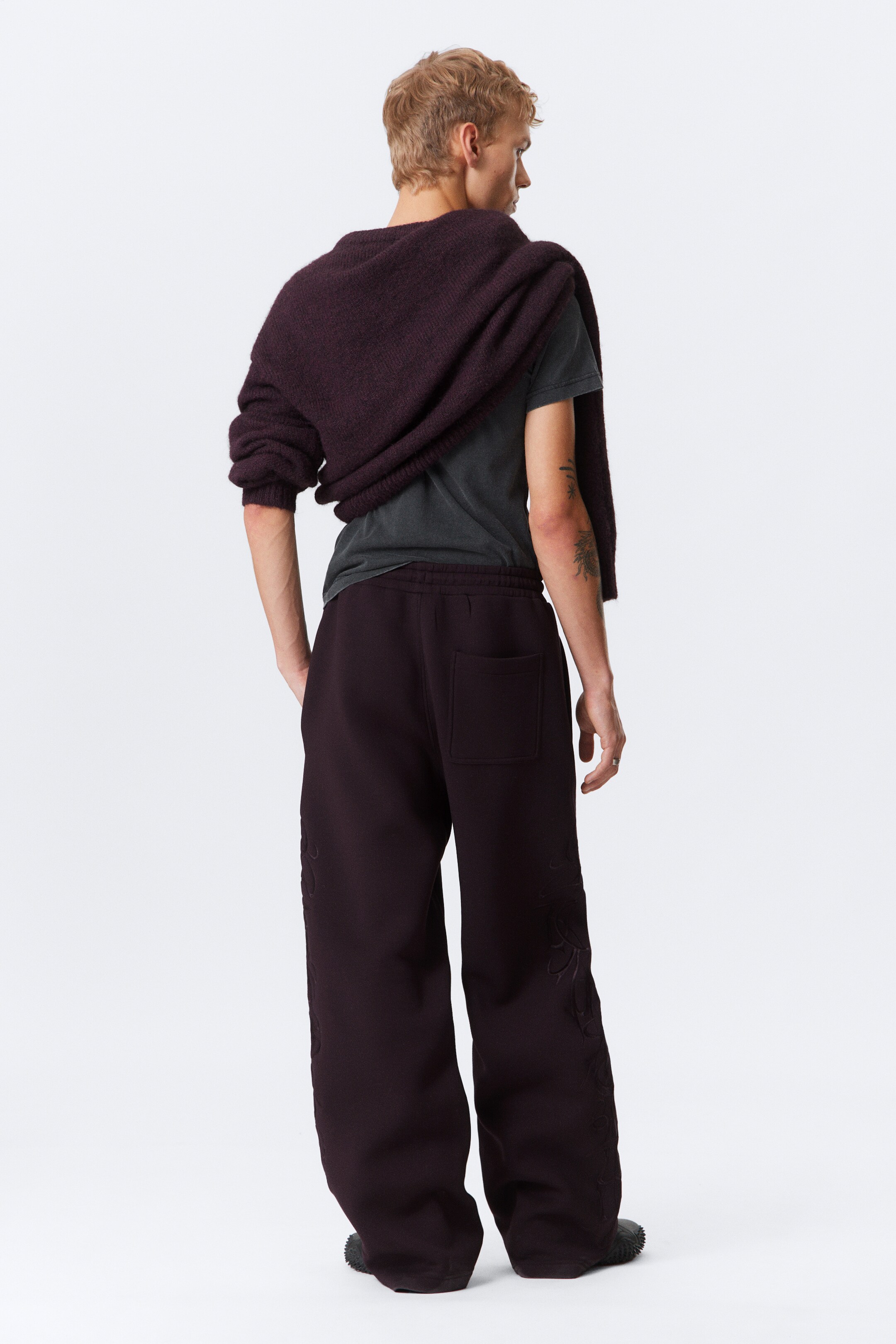Agrandir l'image: Un pantalon ample de couleur bordeaux foncée est porté, vu de dos et légèrement vers la droite. Le pantalon présente une seule poche plaquée à l'arrière droit et une broderie subtile, ton sur ton, sur les deux jambes inférieures.