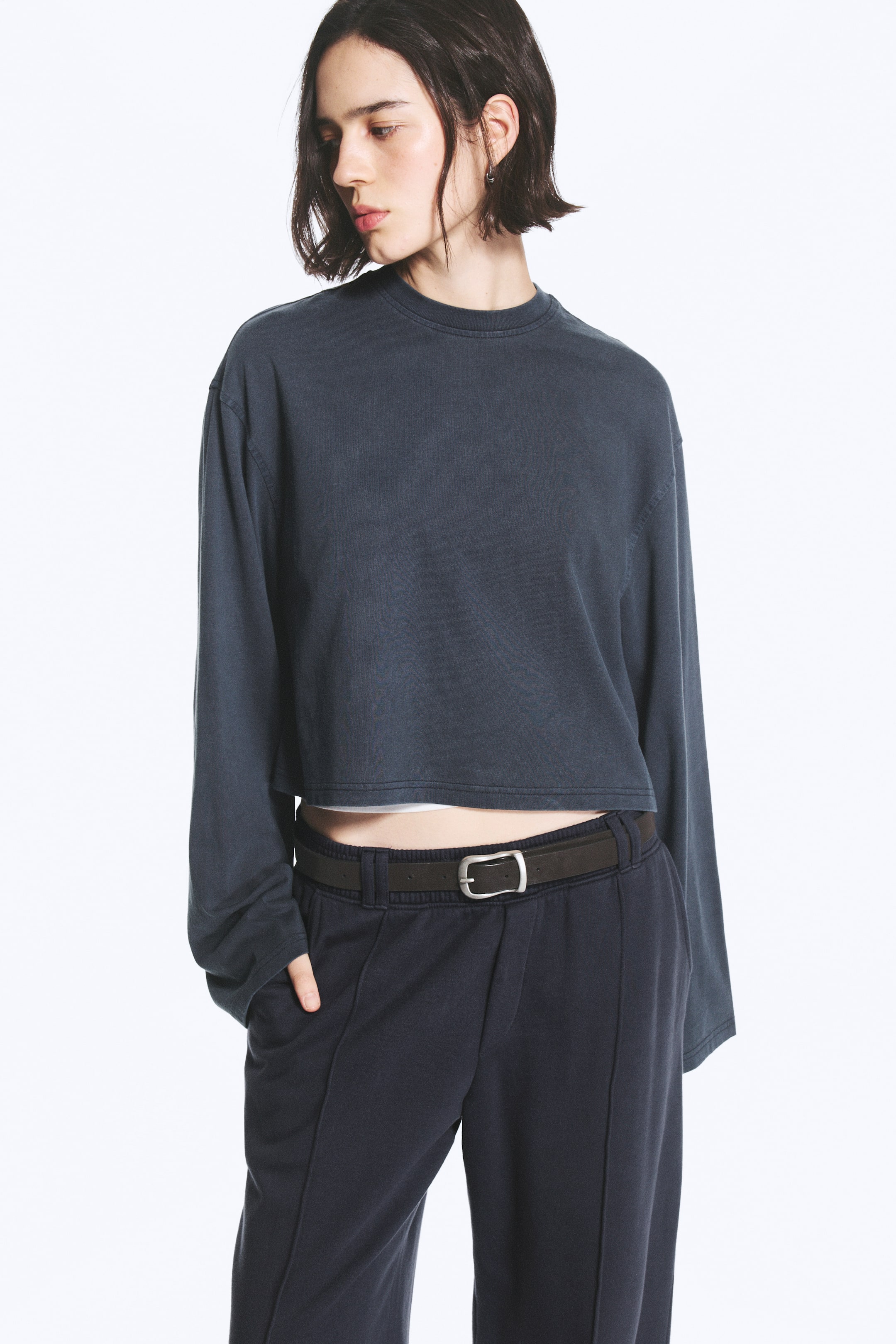 Agrandir l'image: Le mannequin porte un T-shirt court à manches longues, gris foncé, avec un col rond et des épaules tombantes. Ce haut ample et carré est superposé sur une couche blanche en dessous et associé à un pantalon foncé.