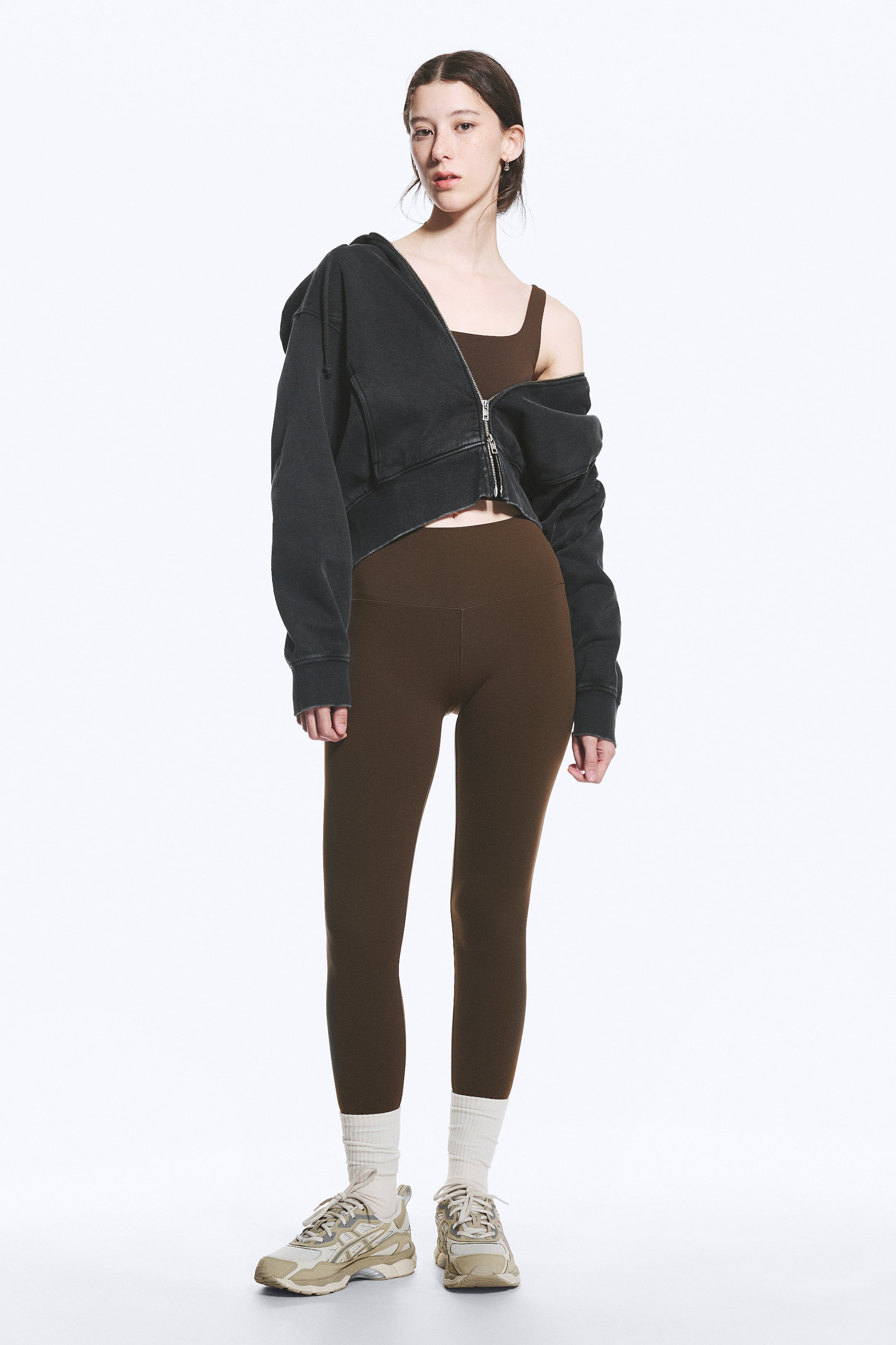 Größeres Bild ansehen: Ein verblasstes, dunkelgraues, kurzes Kapuzen-Sweatshirt mit Reißverschluss wird teilweise von den Schultern getragen, wodurch ein braunes Crop-Top mit quadratischem Ausschnitt darunter sichtbar wird. Hoch taillierte braune Leggings ergänzen das Top und reichen bis zu weißen, gerippten Crew-Socken und beigefarbenen Sportsneakern.