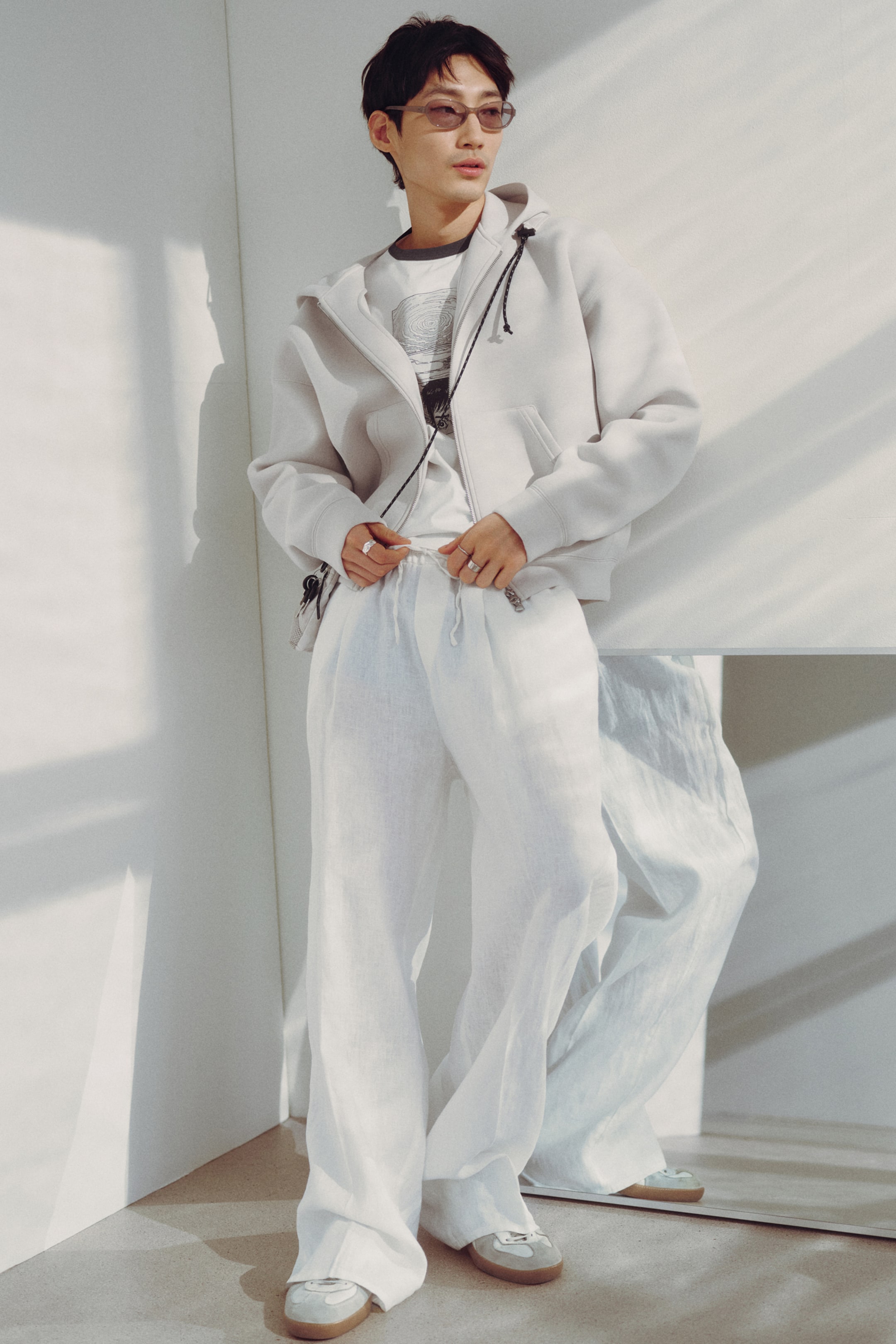 Visualizza immagine più grande: Una felpa con cappuccio a zip intera di colore grigio chiaro viene indossata, leggermente inclinata a destra per mostrare la sua vestibilità rilassata, caratterizzata da un cappuccio con coulisse, tasche frontali e polsini e orlo a coste, ed è stratificata sopra una t-shirt grafica.