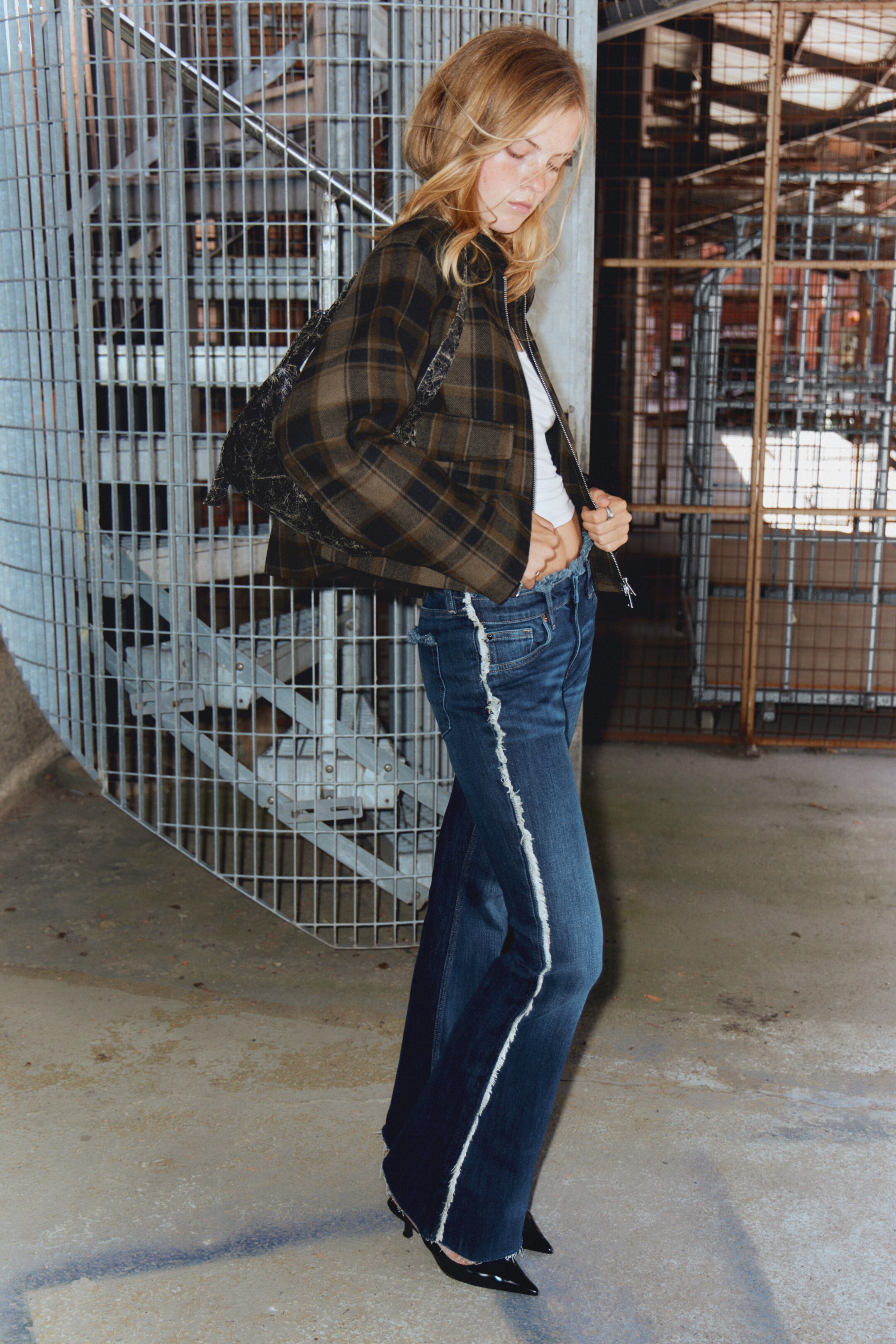 Agrandir l'image: Une veste courte à carreaux olive foncé et noire, avec une fermeture éclair à l'avant, est portée sur un haut blanc. Un jean large en denim délavé foncé, distingué par une bande effilochée blanche courant le long de chaque couture extérieure, est porté avec des talons noirs à bout pointu.