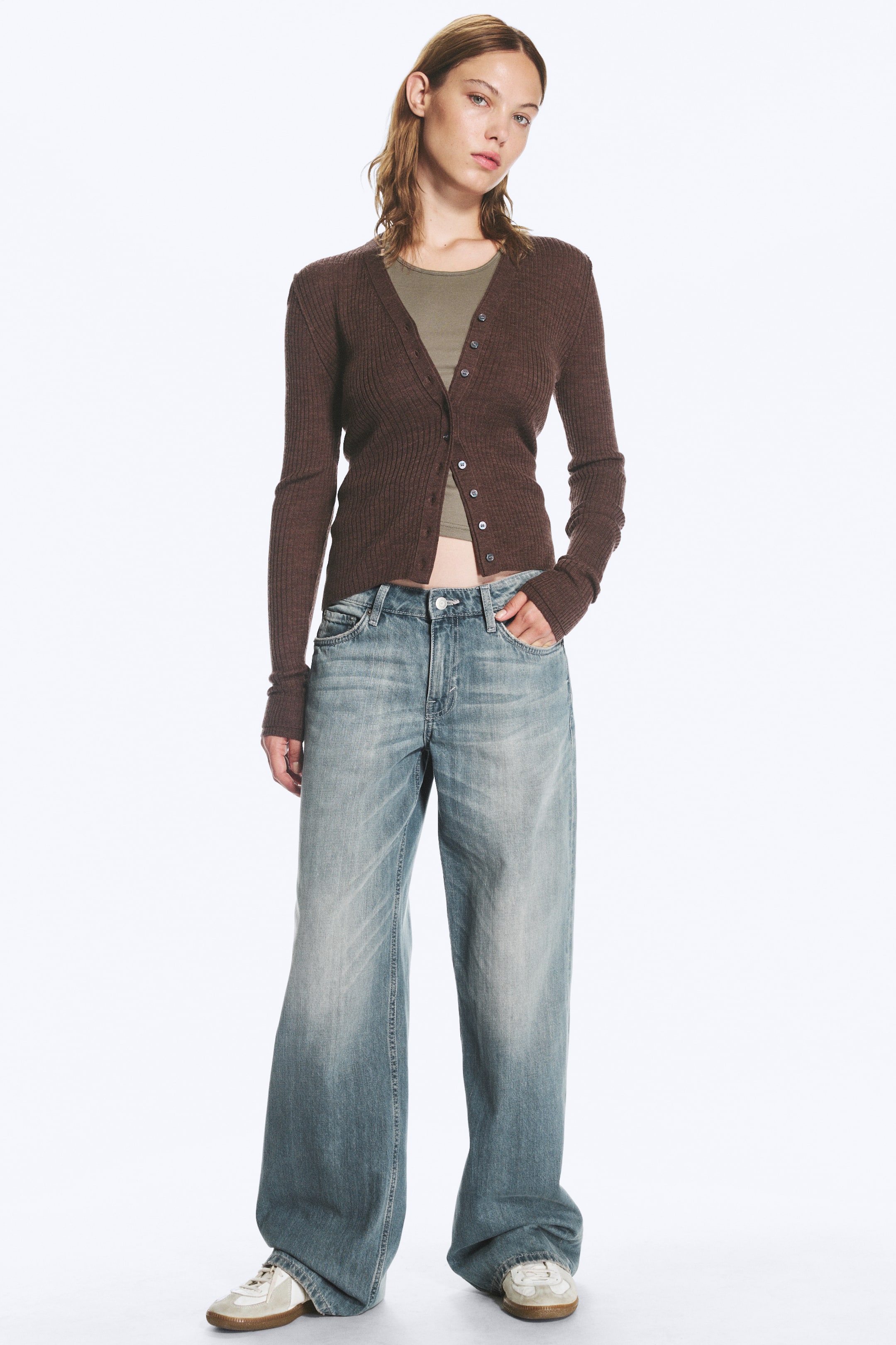 Agrandir l'image: Une paire de jeans taille basse à jambe large, dans un délavé bleu vieilli, est portée par le mannequin, qui se tient debout avec une main glissée dans une poche avant. Ce jean long et ample est associé à un cardigan côtelé marron, un haut vert olive et des baskets plates.