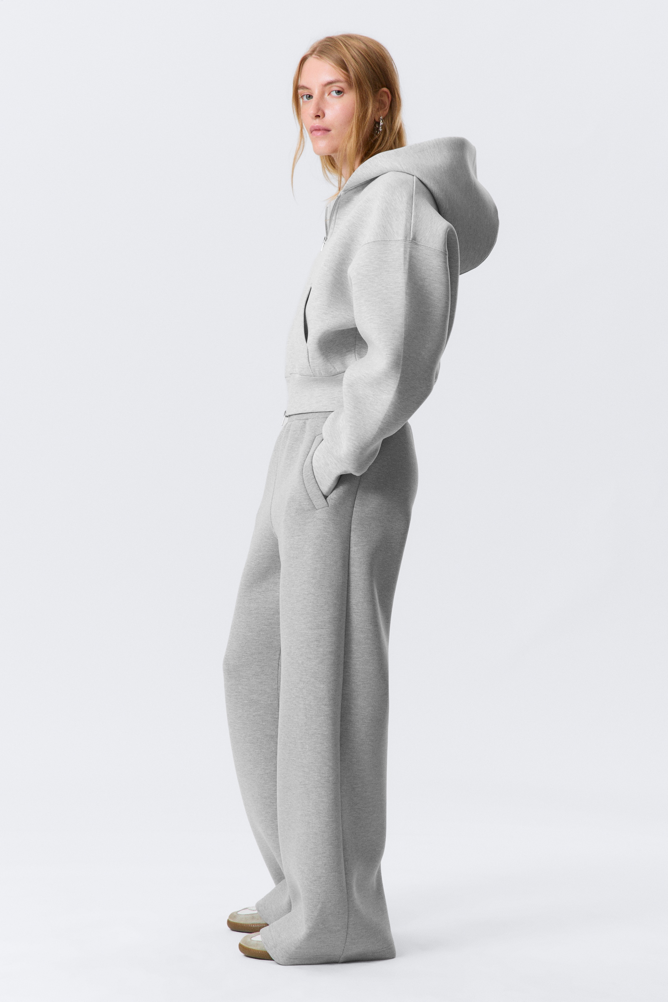 Agrandir l'image: Le survêtement gris, présenté de profil face à droite, comprend un hoodie court à fermeture éclair complète avec une capuche et des épaules tombantes, ainsi qu'un pantalon de jogging à jambes larges assorti avec des poches latérales.