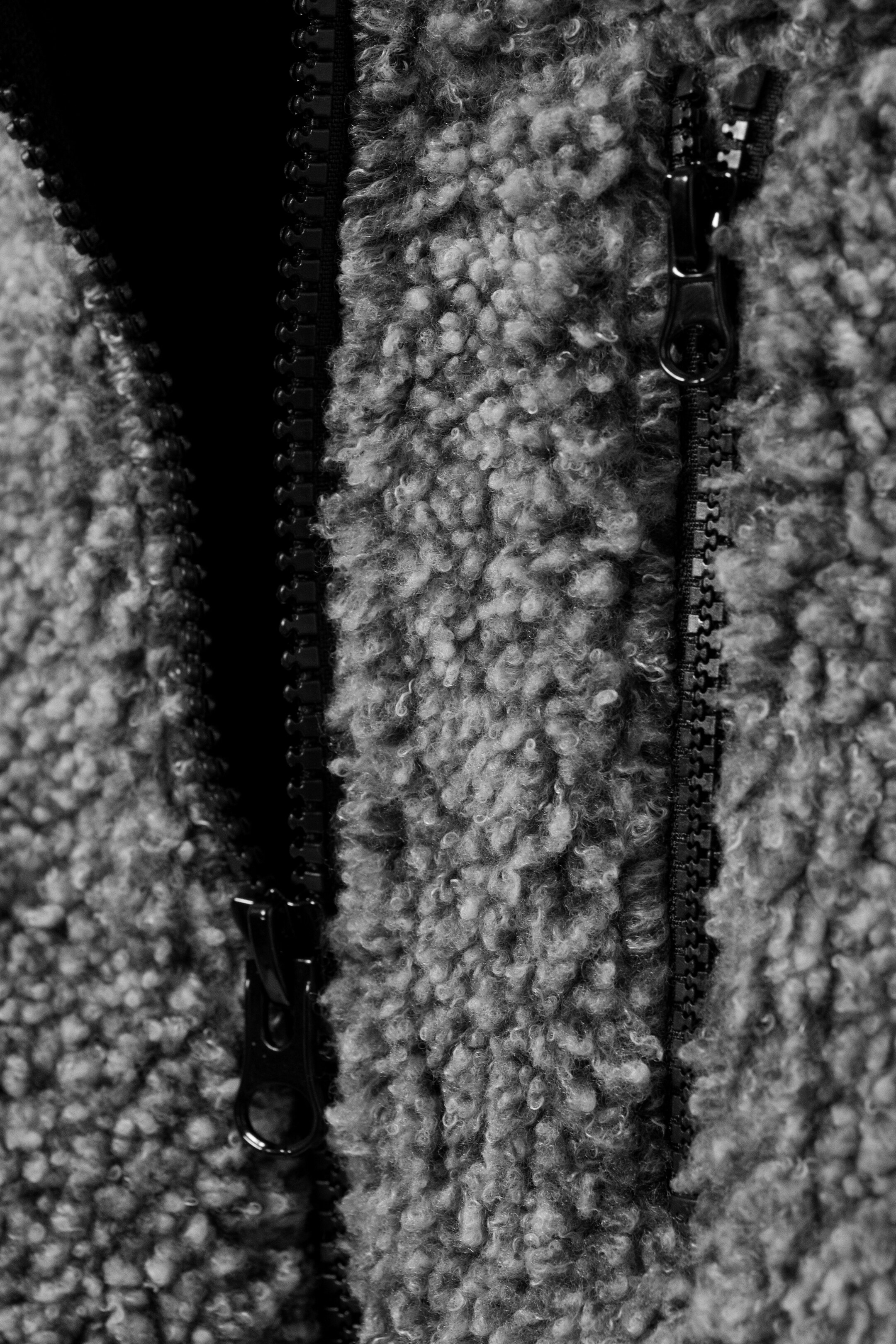 Agrandir l'image: Un vêtement en polaire teddy gris présente une texture douce et bouclée avec deux fermetures éclair noires, l'une partiellement ouverte révélant une couche intérieure sombre, et l'autre fermée avec son tirette visible.