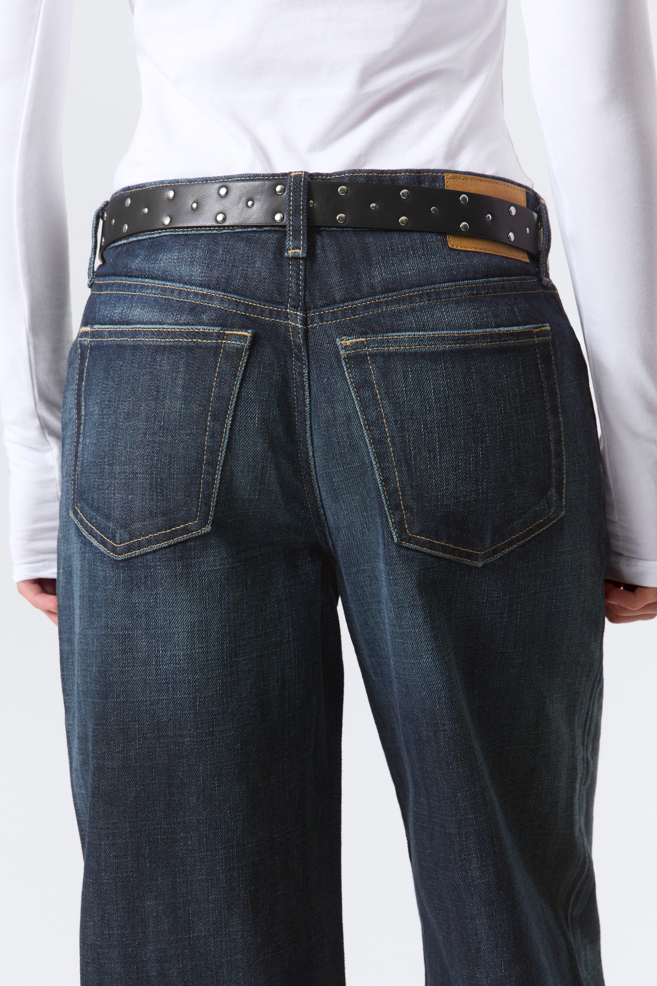 View larger image: Dark wash denim jeans are being presented from the back, featuring two patch pockets with contrast stitching. A black studded belt is threaded through the belt loops, with a light brown leather patch visible at the back waistband.
