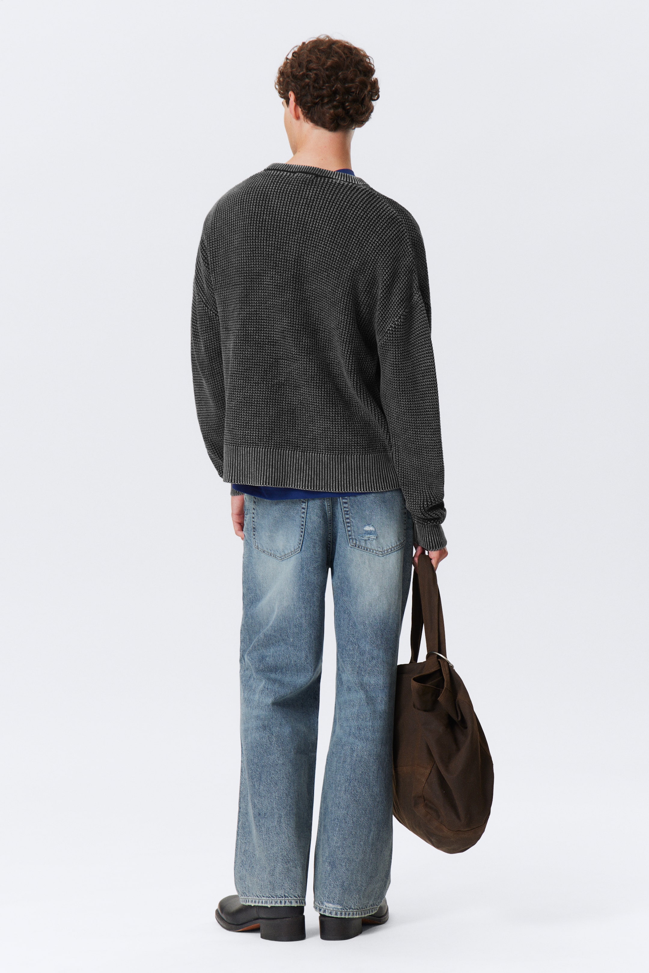 Größeres Bild anzeigen: Ein dunkelgrauer Waffelstrickpullover mit gerippten Bündchen und Saum wird von hinten präsentiert, über einem blauen Oberteil geschichtet. Hell gewaschene, weit geschnittene Jeans mit abgenutzten Details werden ebenfalls getragen und mit schwarzen Stiefeln kombiniert. Eine dunkelbraune, lässige Tragetasche mit einem Metalldetail wird gehalten.