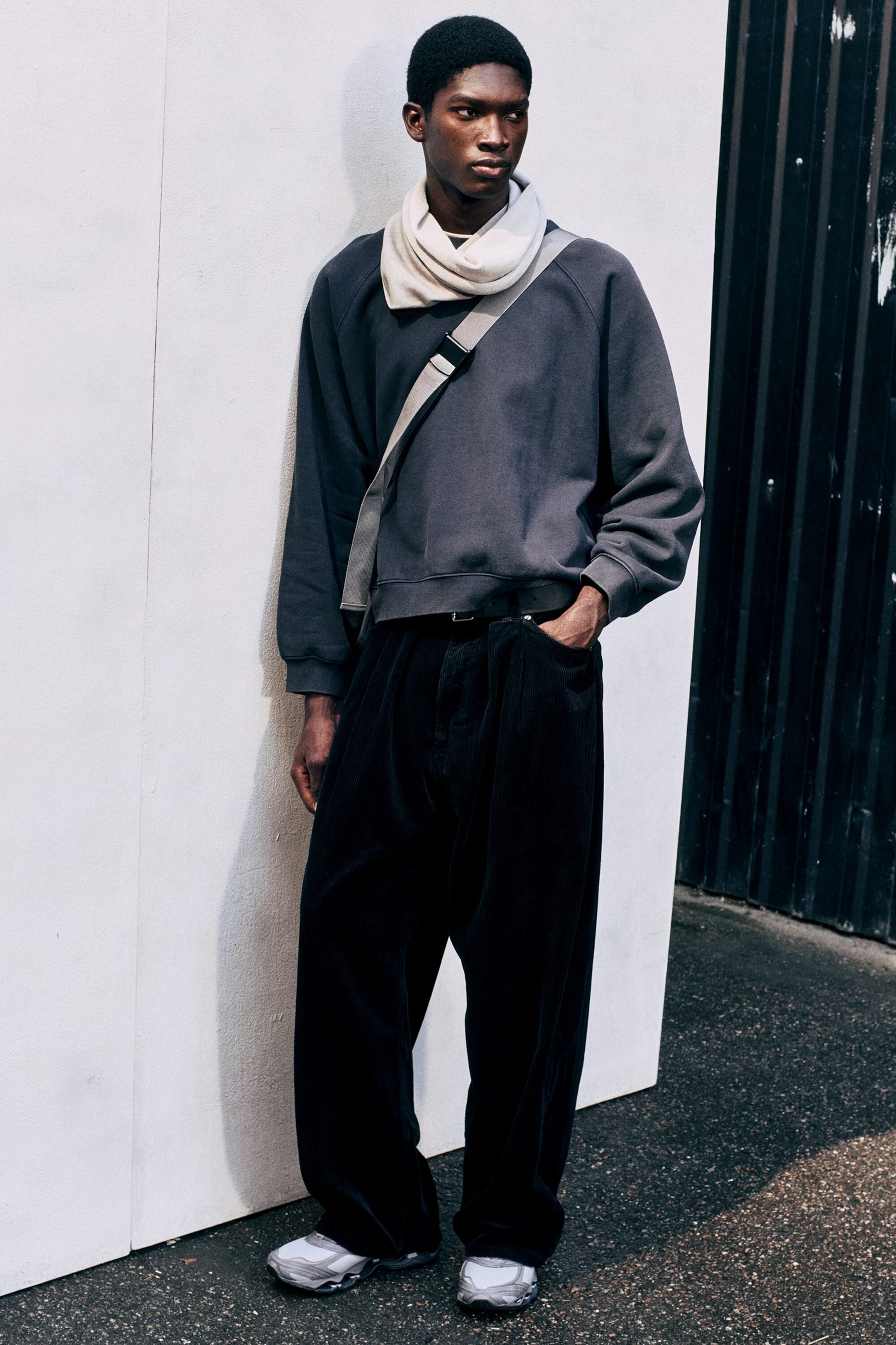 Ver imagen más grande: Un pantalón de pana oscuro de corte holgado, con pinzas delanteras y silueta baggy, lo luce la modelo, que está de pie con una mano en el bolsillo. El pantalón se combina con una sudadera gris de manga larga, un cuello alto de color crema y unas zapatillas grises.