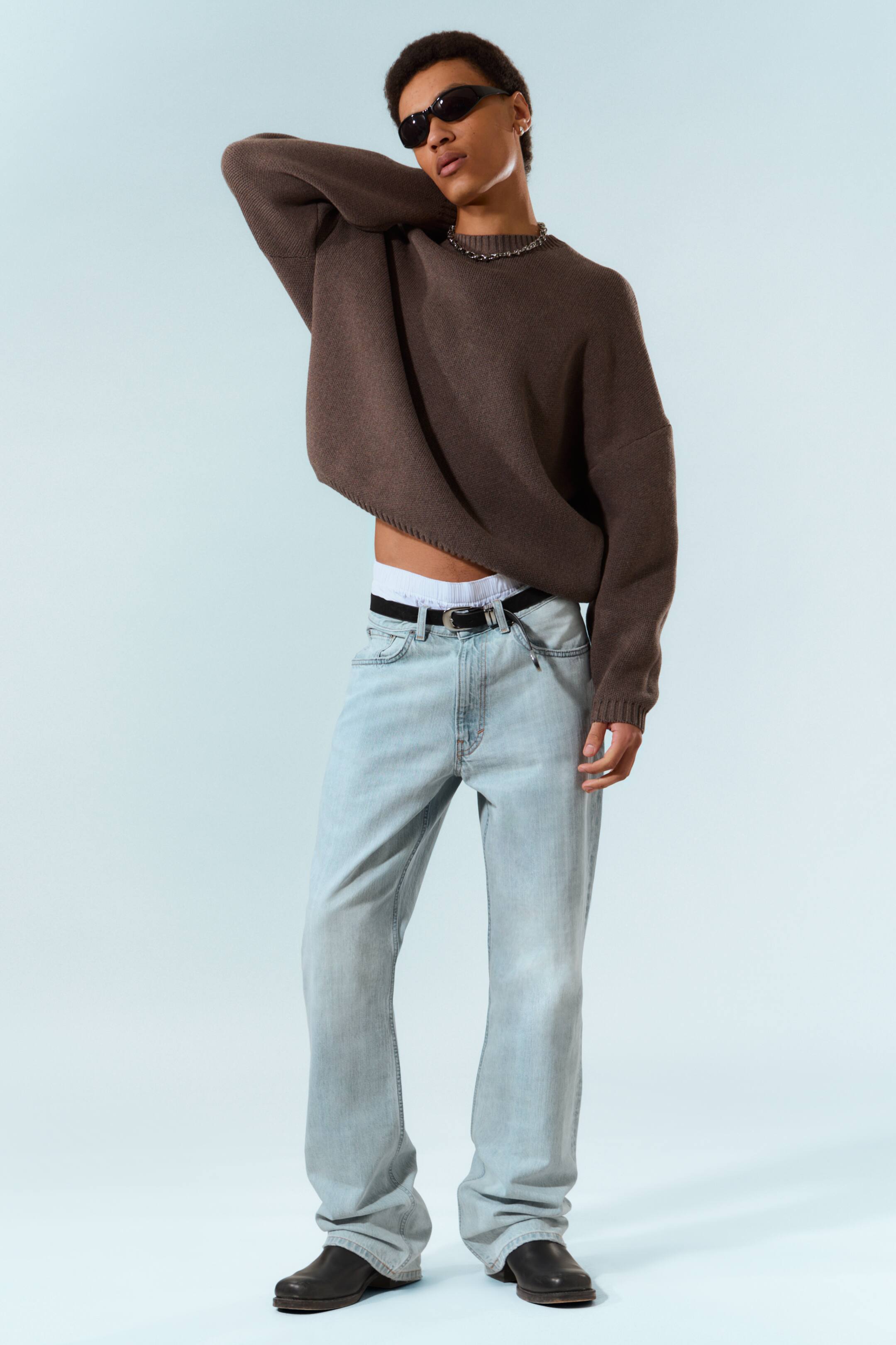 Grotere afbeelding bekijken: Een oversized donkerbruine trui met ronde hals, met lange losse mouwen, wordt gedragen over licht gewassen, relaxed-fit jeans die vastgehouden worden door een zwarte riem. Witte onderkleding piept boven de jeans uit, een zilverkleurige ketting siert de ronde halslijn van de trui, en zwarte chunky laarzen zijn zichtbaar aan de voeten.
