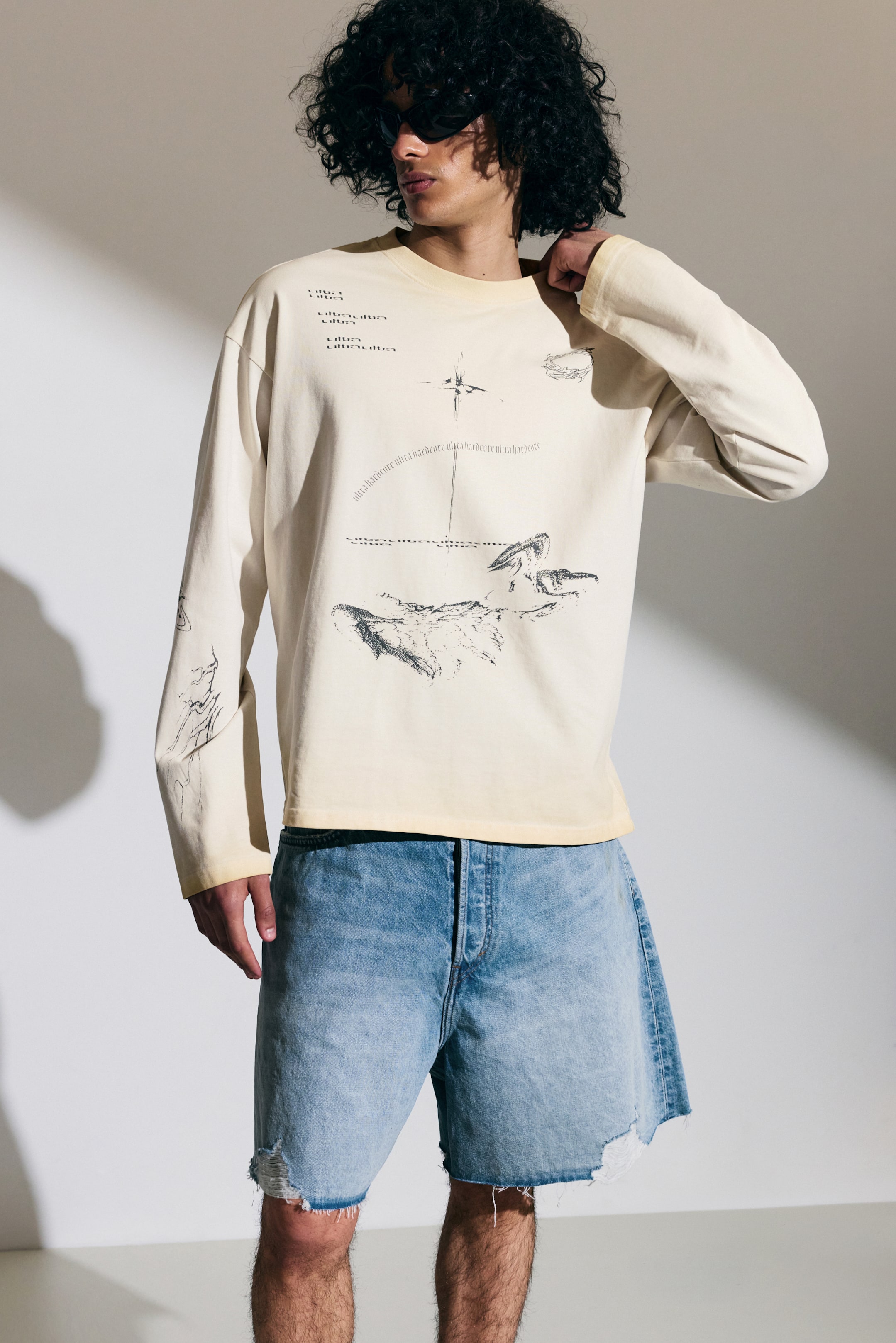 Größeres Bild ansehen: Das cremefarbene Langarm-T-Shirt bietet eine lockere Passform und einen Rundhalsausschnitt. Auf der Vorderseite sind schwarze Grafikdrucke zu sehen, darunter ein kompassähnlicher Sternburst, wiederholte Texte und abstrakte Landschaftsillustrationen, mit zusätzlichen abstrakten Linienzeichnungen, die den linken Ärmel bedecken.