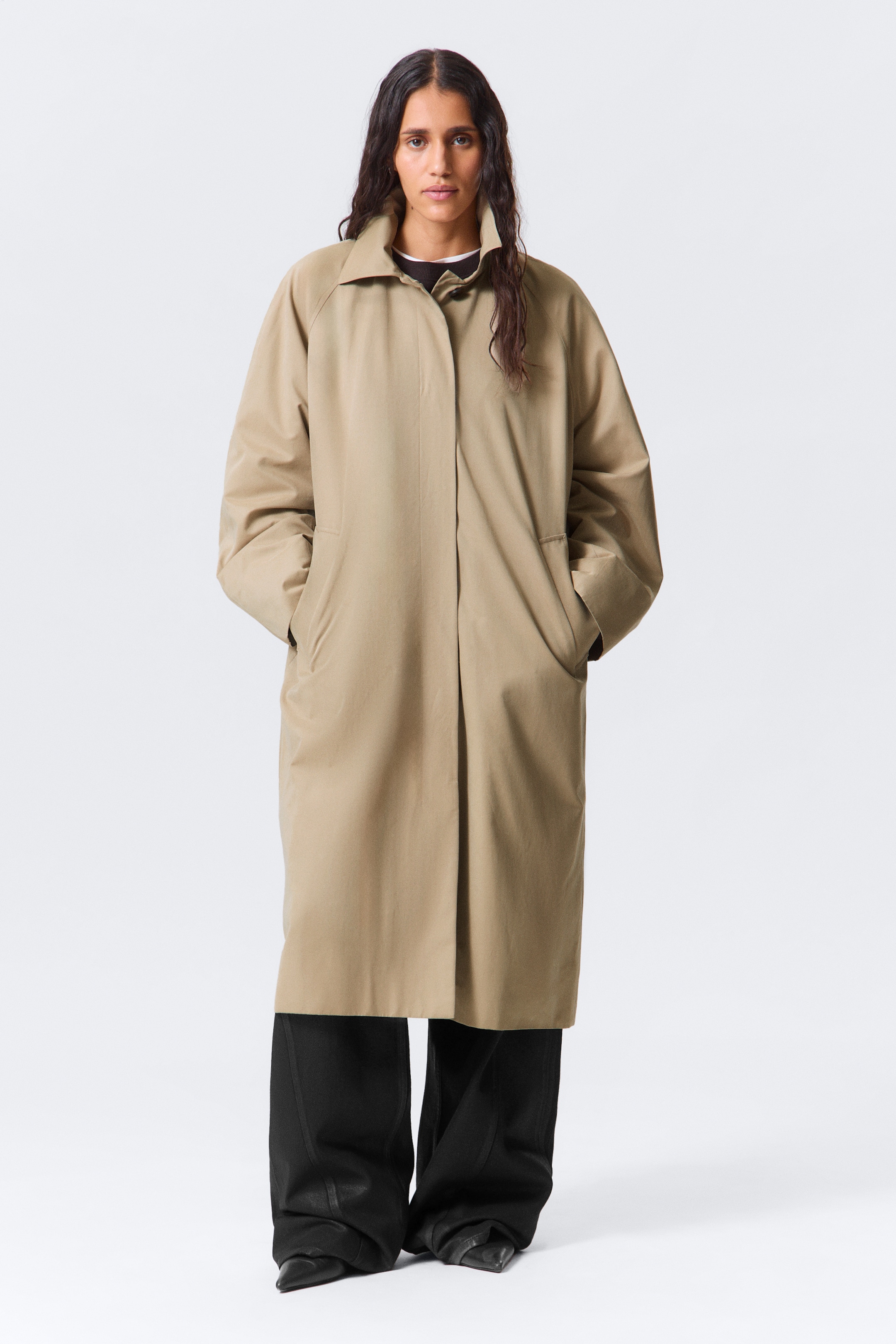 Grotere afbeelding bekijken: Een khaki trenchcoat die naar voren is gericht, wordt gepresenteerd met een klassieke spreidkraag, raglanmouwen en een verborgen knopenlijst. De lange jas heeft een ontspannen, oversized pasvorm met verticale paspelzakken, strekt zich uit voorbij de knieën en is gelaagd over zwarte breedpijpbroeken, met een donkere top zichtbaar bij de halslijn.