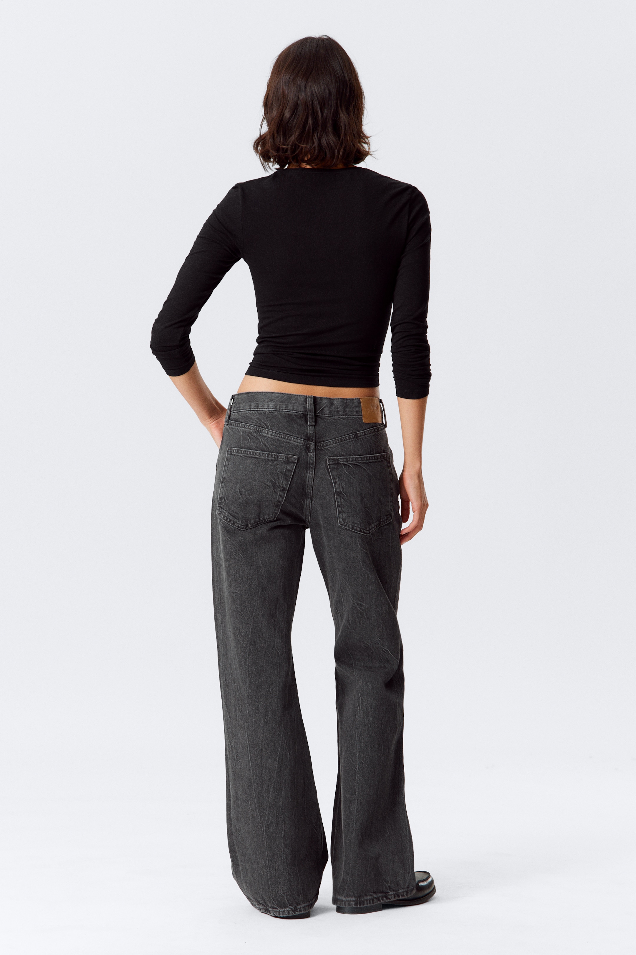 Grösseres Bild ansehen: Eine Person trägt ein schwarzes langärmliges Oberteil und dunkel gewaschene Jeans mit weitem Bein, von hinten betrachtet. Das enganliegende Oberteil ist bauchfrei und zeigt den Bund der locker sitzenden Jeans, die eine verblasste dunkle Waschung, zwei aufgesetzte Gesäßtaschen und einen hellbraunen Leder-Markenaufnäher aufweisen.