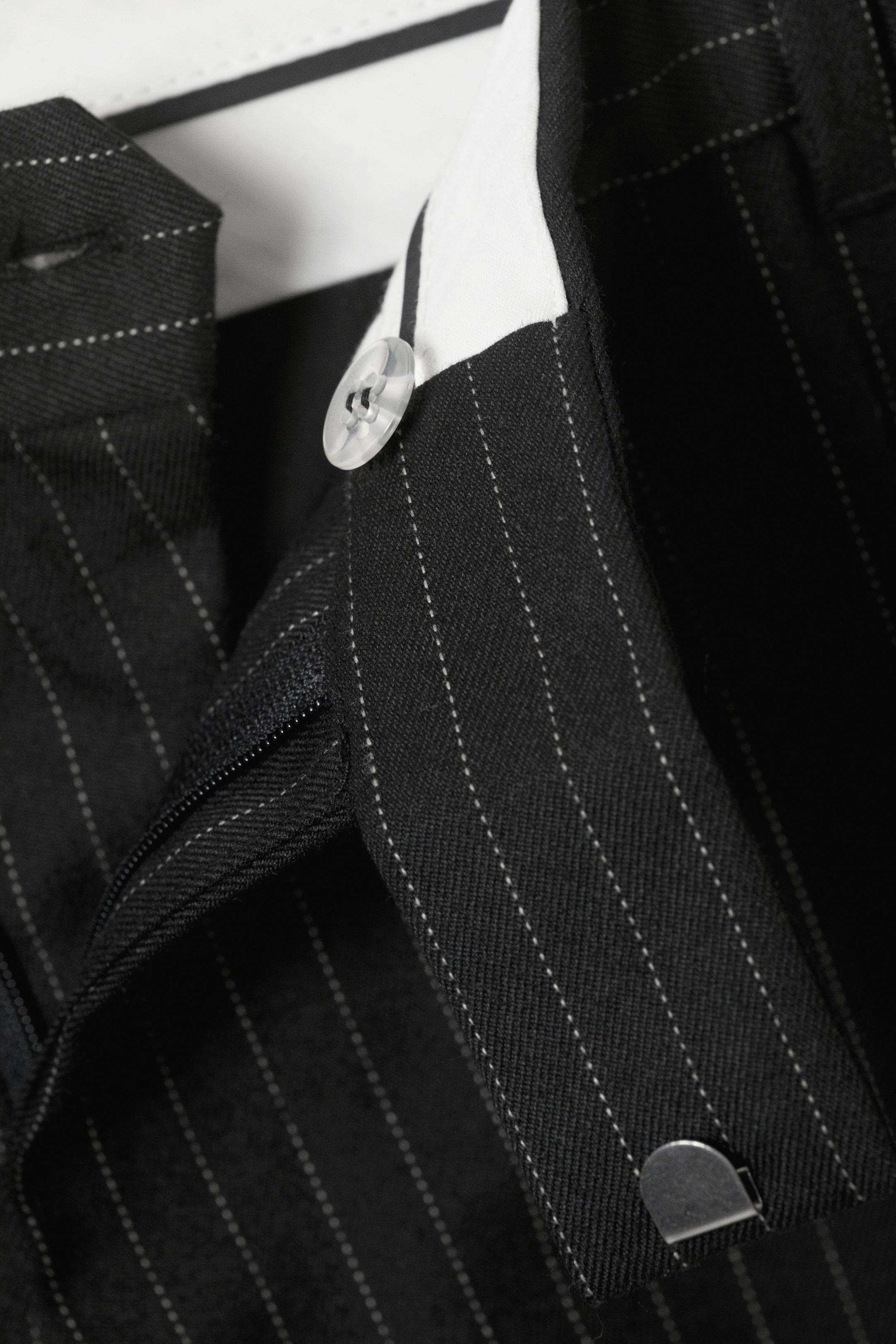 Agrandir l'image: Un vêtement à rayures noires met en avant les détails de sa fermeture frontale, y compris un bouton transparent à quatre trous fixant un rabat, une fermeture éclair visible et une attache en métal à crochet et barre sécurisant la ceinture.
