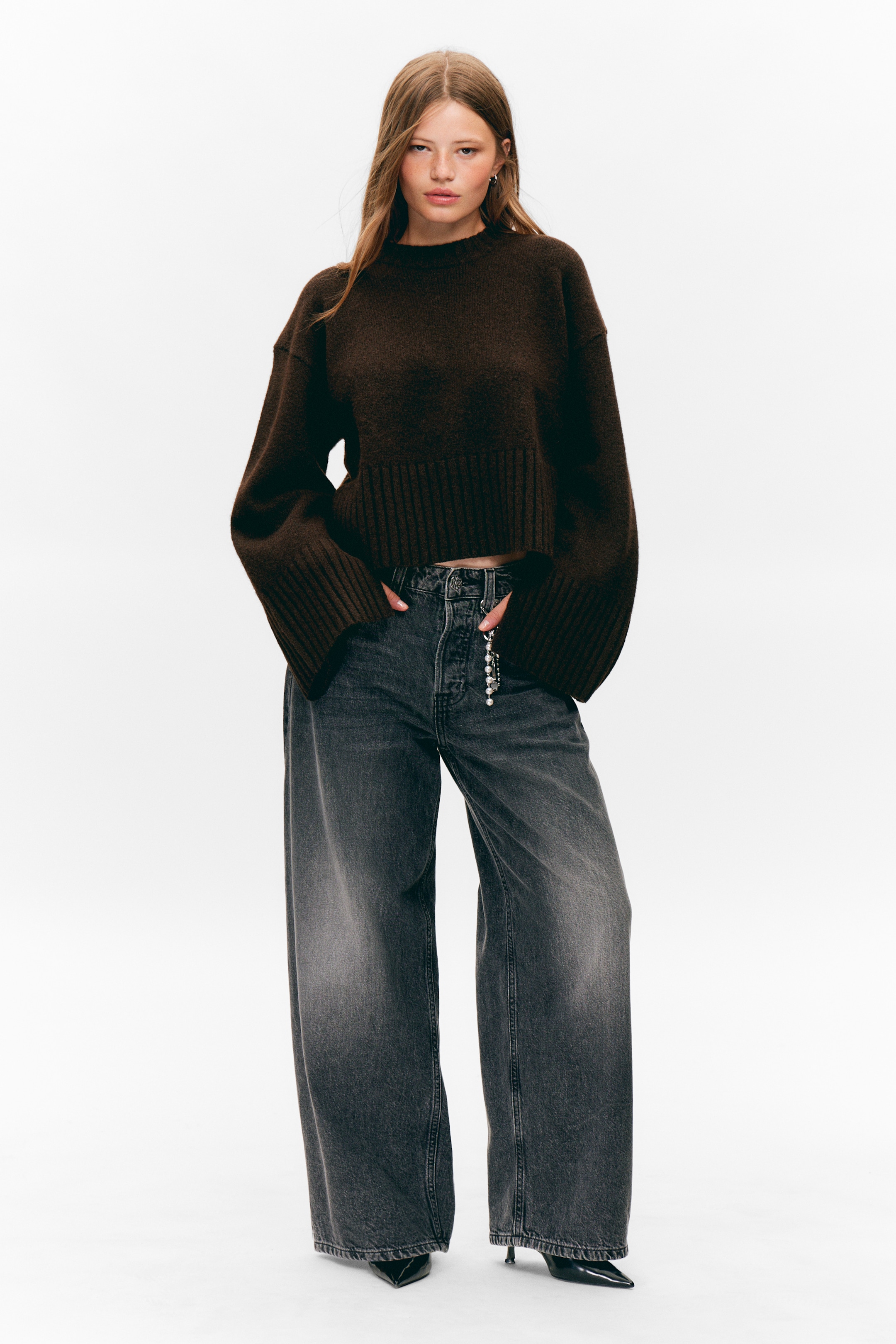 Ingrandisci l'immagine: Un maglione corto marrone scuro, caratterizzato da un girocollo, maniche ampie con polsini a coste profonde e un orlo a coste profonde, è indossato. È abbinato a jeans grigio scuro a vita alta e gamba larga che mostrano una chiusura a bottoni visibile e un accessorio a catena con perline di perle appeso a un passante per cintura.