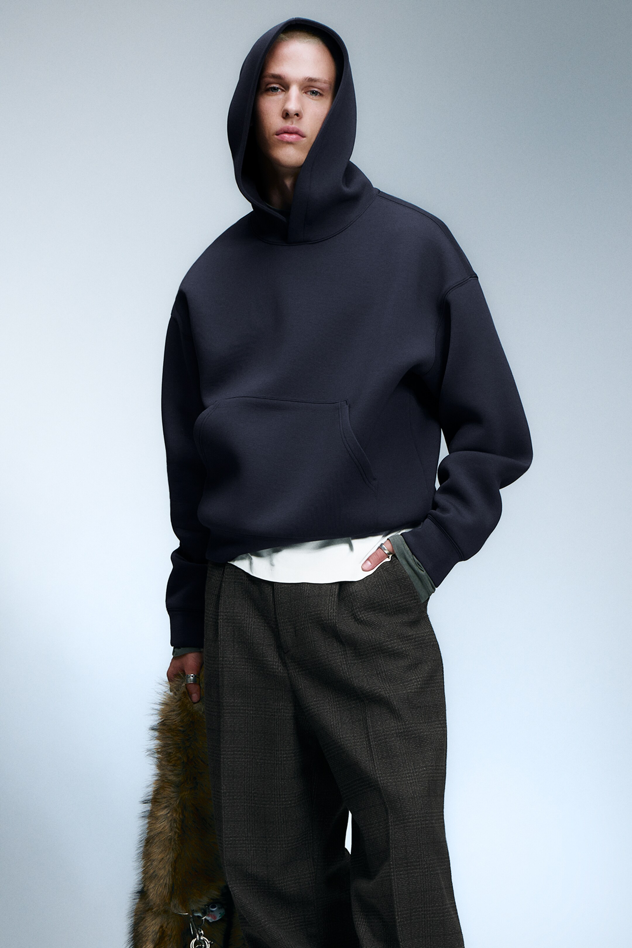 Grotere afbeelding bekijken: Een donkerblauwe hoodie, gemaakt van een gladde, gestructureerde stof, wordt gepresenteerd vanuit een frontale hoek, met een prominente capuchon en een grote kangoeroezak. Een wit t-shirt is zichtbaar onder de zoom gelaagd, met groene lange mouwen die bij de manchetten zichtbaar zijn.