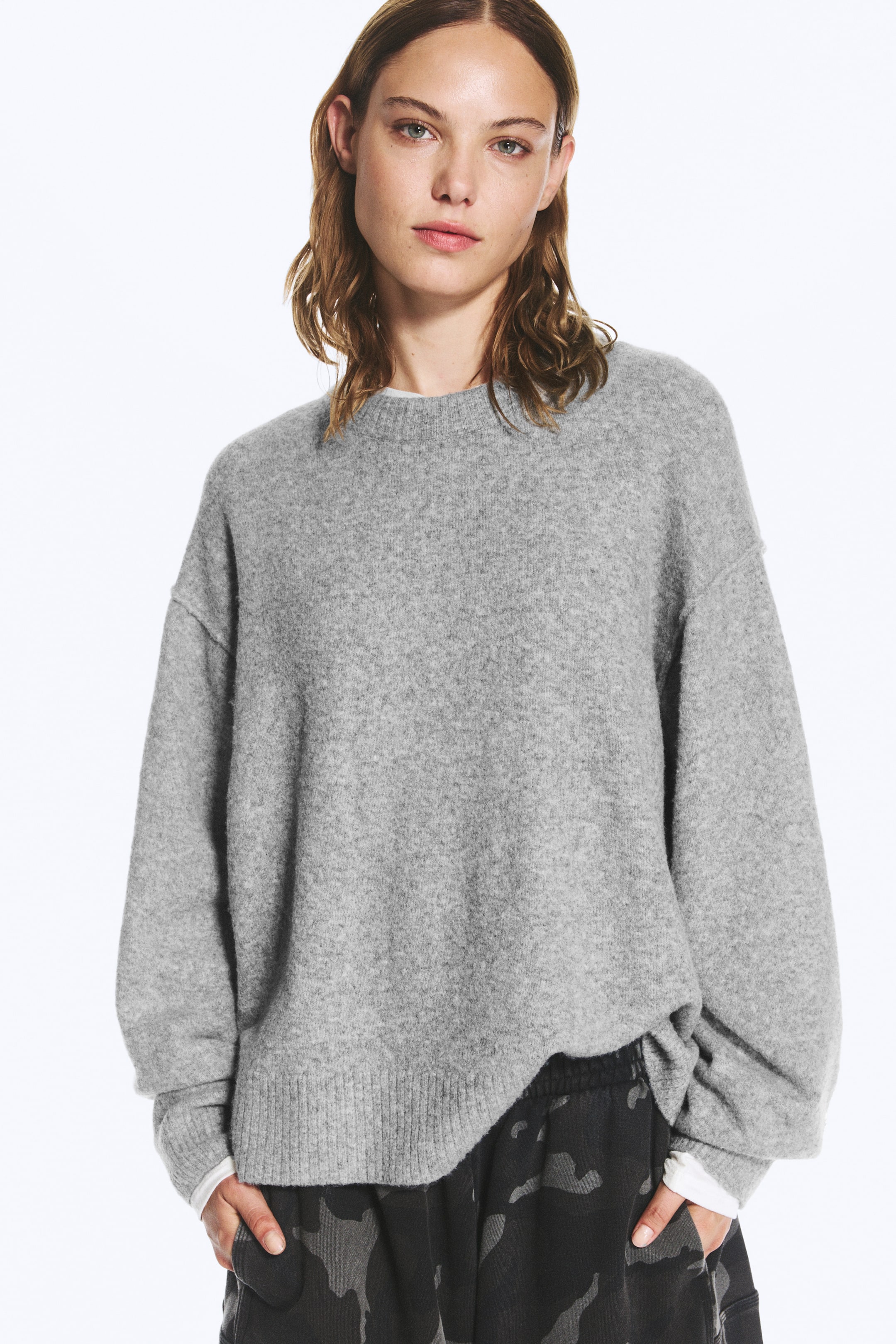 Agrandir l'image: Un pull en maille gris clair à coupe décontractée est porté par le mannequin, avec un col rond côtelé, des épaules tombantes et des poignets côtelés. Le pull est superposé à un sous-pull blanc et associé à un pantalon foncé à motifs.