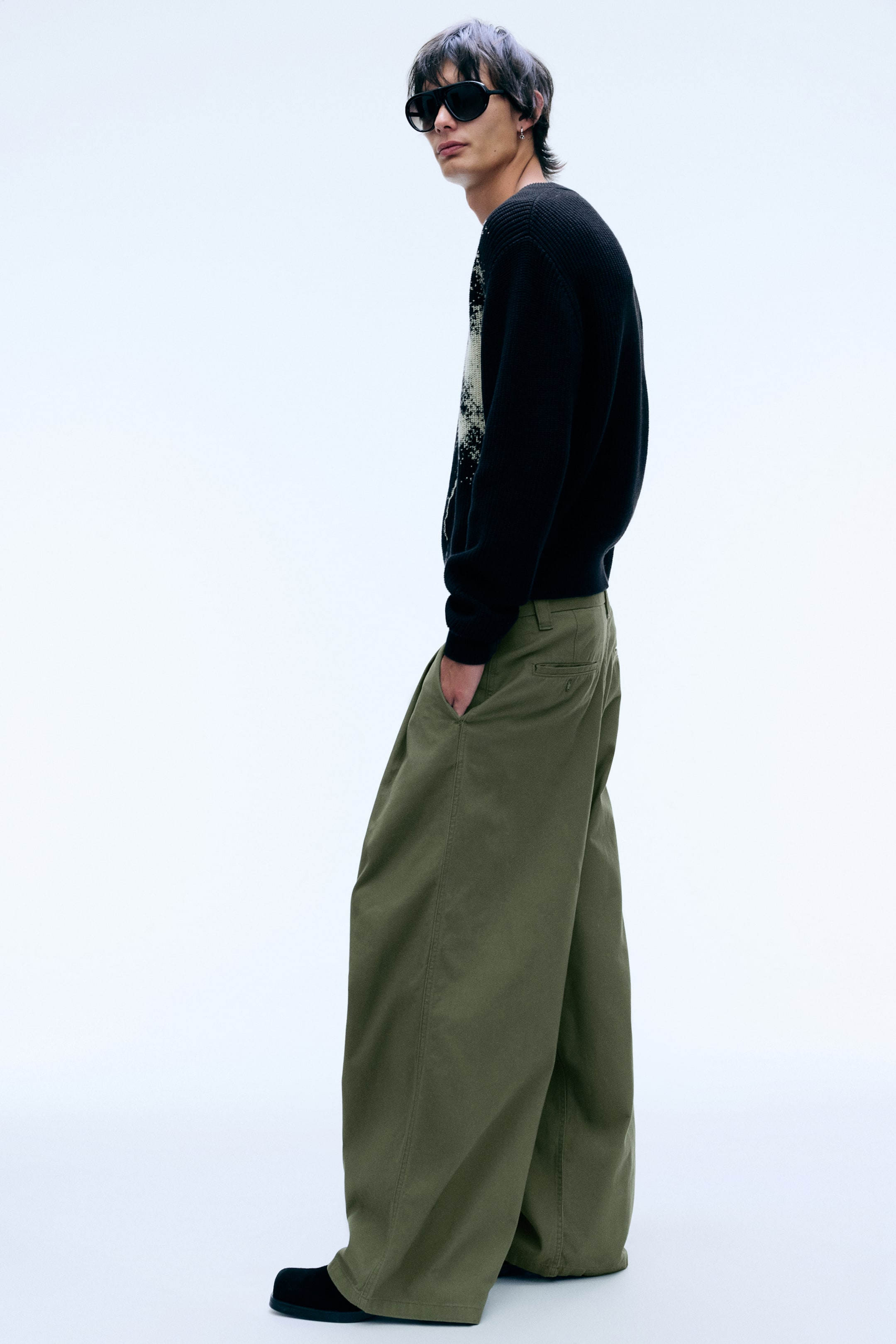 Visualizza l'immagine ingrandita: Pantaloni chino loose fit verde oliva indossati dalla modella, in posa di profilo con una mano appoggiata in una tasca laterale obliqua. I pantaloni a gamba ampia presentano una silhouette baggy con pieghe sul davanti e una tasca a filetto posteriore, e sono abbinati a un maglione in maglia nero e scarpe nere.