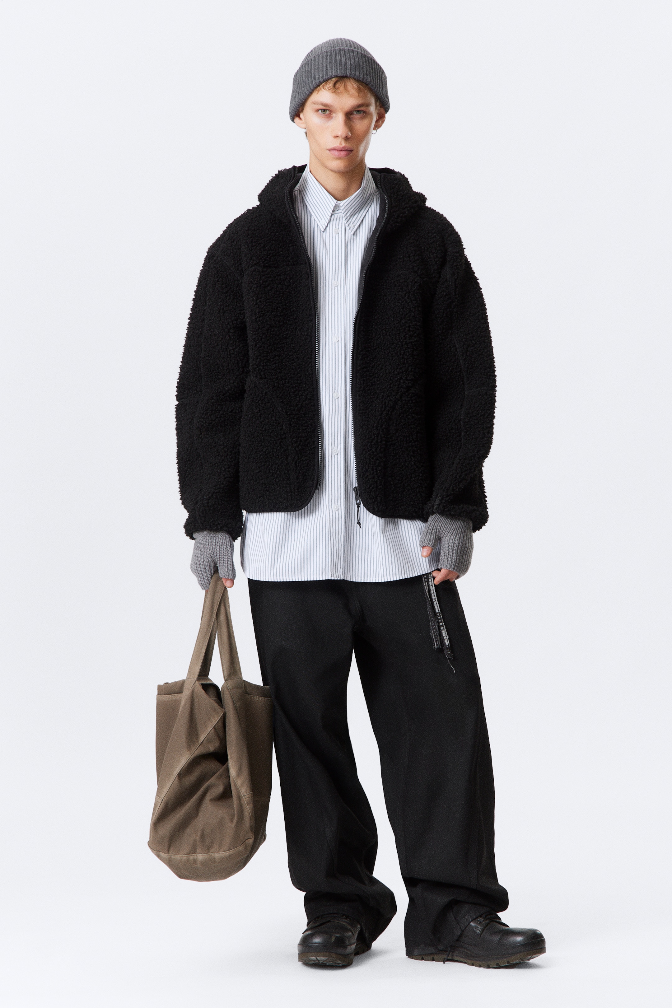 Visualizza immagine più grande: Una giacca in pile sherpa con cappuccio nero viene presentata, rivolta in avanti, con una chiusura frontale a tutta zip. È caratterizzata da un orlo arrotondato ed è stilizzata aperta su una camicia a righe di colore chiaro.