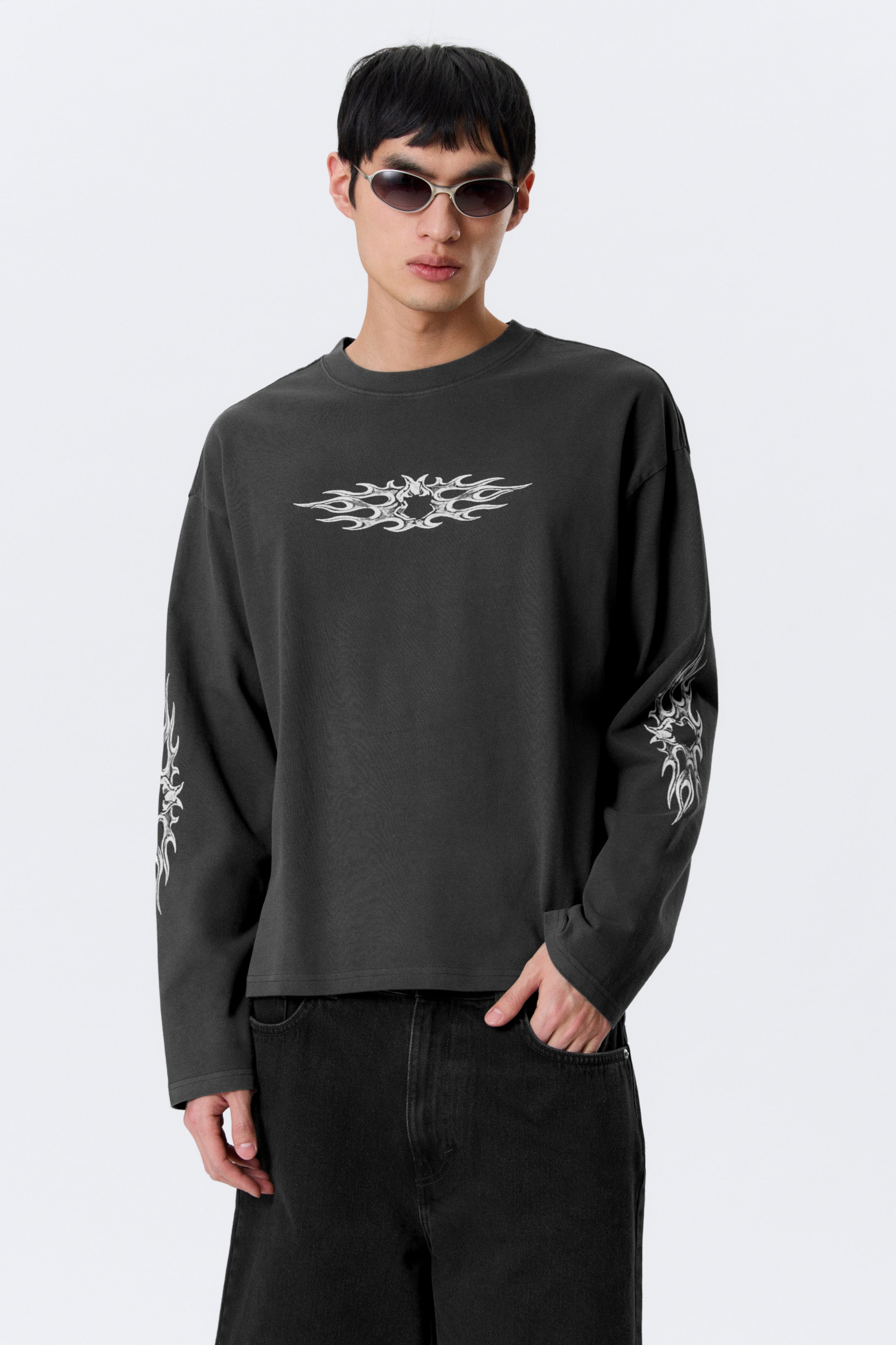 Visa större bild: En mörkgrå långärmad t-shirt med rund hals och avslappnad passform. Den visar upp en stor vit tribalflamgrafik över bröstet och mindre matchande grafik på båda underarmarna.