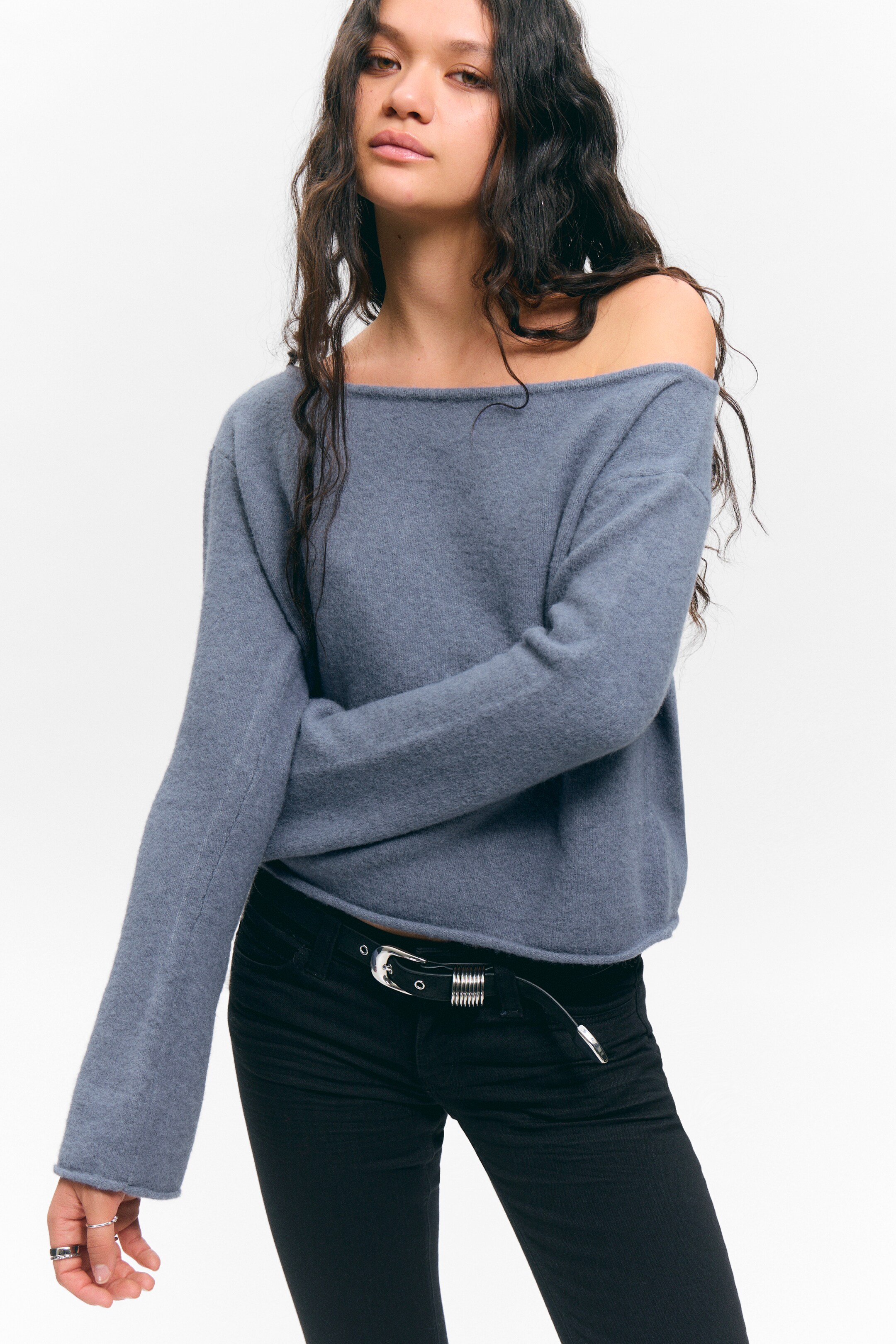 Ingrandisci l'immagine: Un maglione color ardesia a collo a barca viene indossato, presentando una vestibilità rilassata e una texture morbida e soffice. La sua ampia scollatura cade da una spalla, e le maniche lunghe e larghe si estendono fino ai polsi. L'orlo dritto del maglione si appoggia alla vita, sopra pantaloni neri.