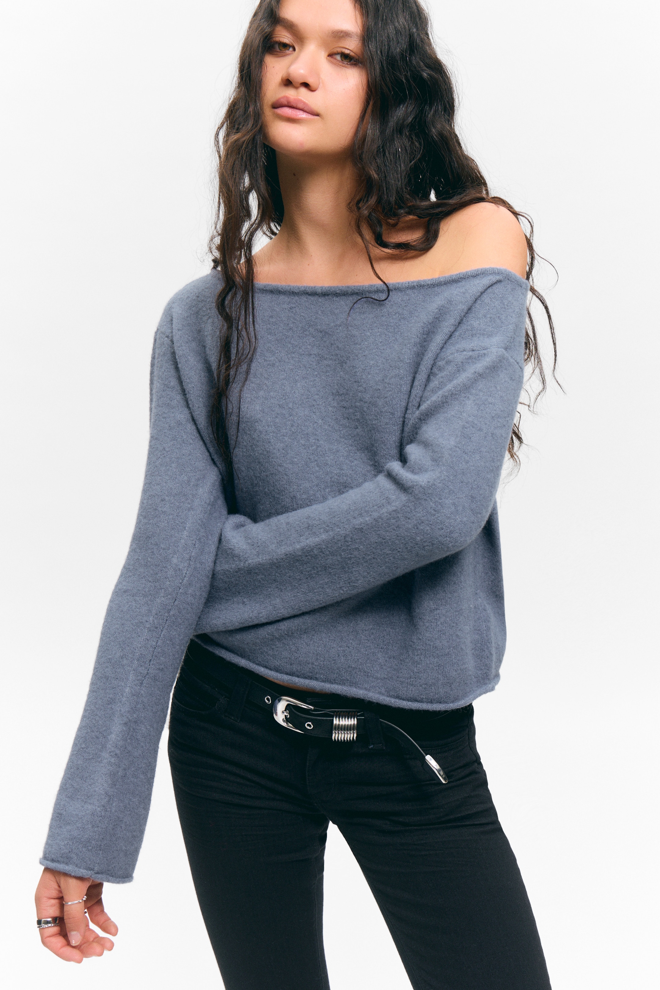 Größeres Bild ansehen: Ein schieferblauer Pullover mit Bootsausschnitt wird getragen, präsentiert eine lockere Passform und eine weiche, flauschige Textur. Sein weiter Ausschnitt rutscht von einer Schulter, und die langen, lockeren Ärmel reichen bis zu den Handgelenken. Der gerade Saum des Pullovers sitzt auf der Taille, über schwarzen Hosen.