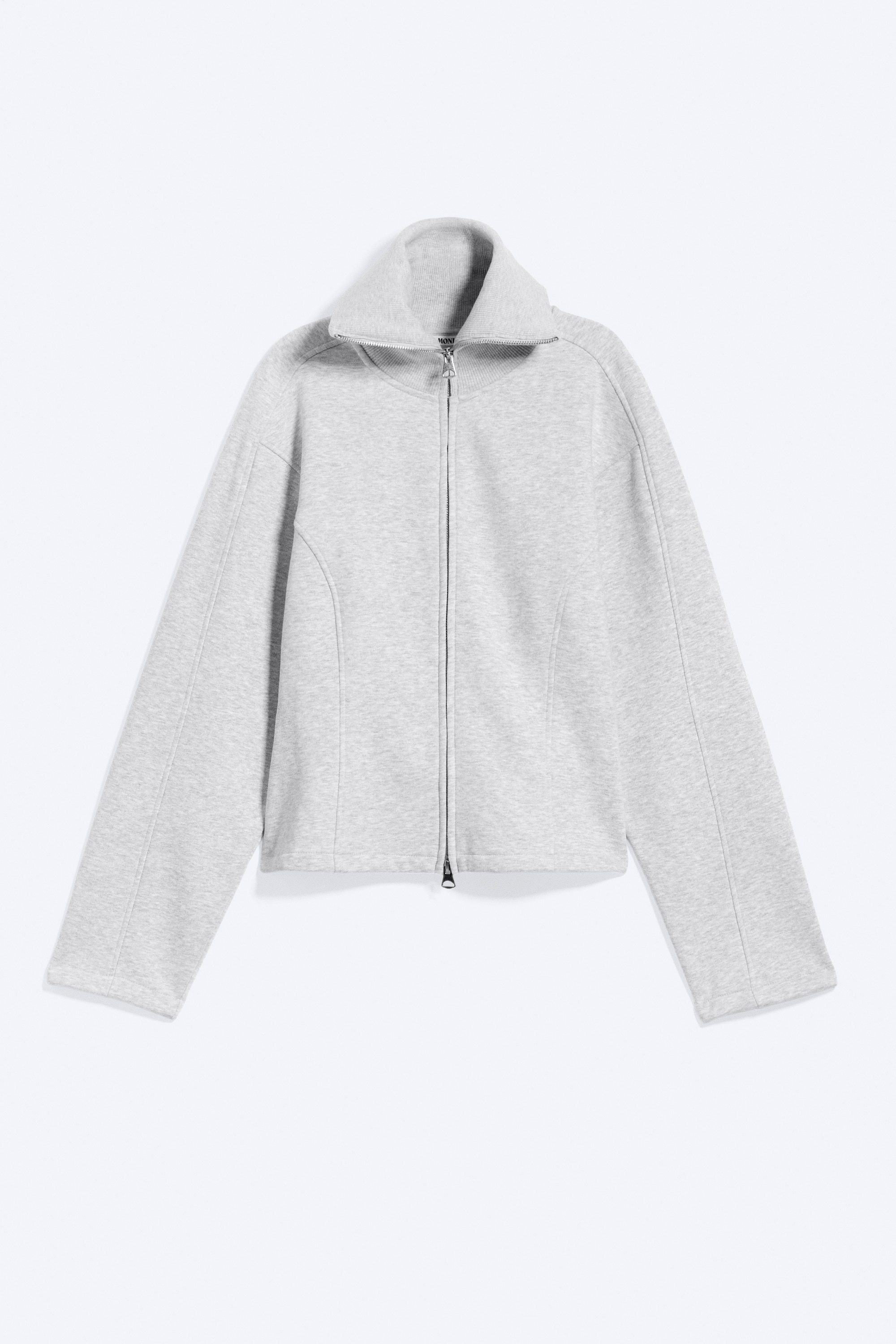 En ljusgrå slim fit-collegetröja med ståkrage och böjda sömsektioner bärs av modellen, som håller i den nedre änden av den delvis öppna tvåvägsdragkedjan. Collegetröjan är stylad över en beige topp och matchad med mörkgrå, texturerade byxor med vida ben.