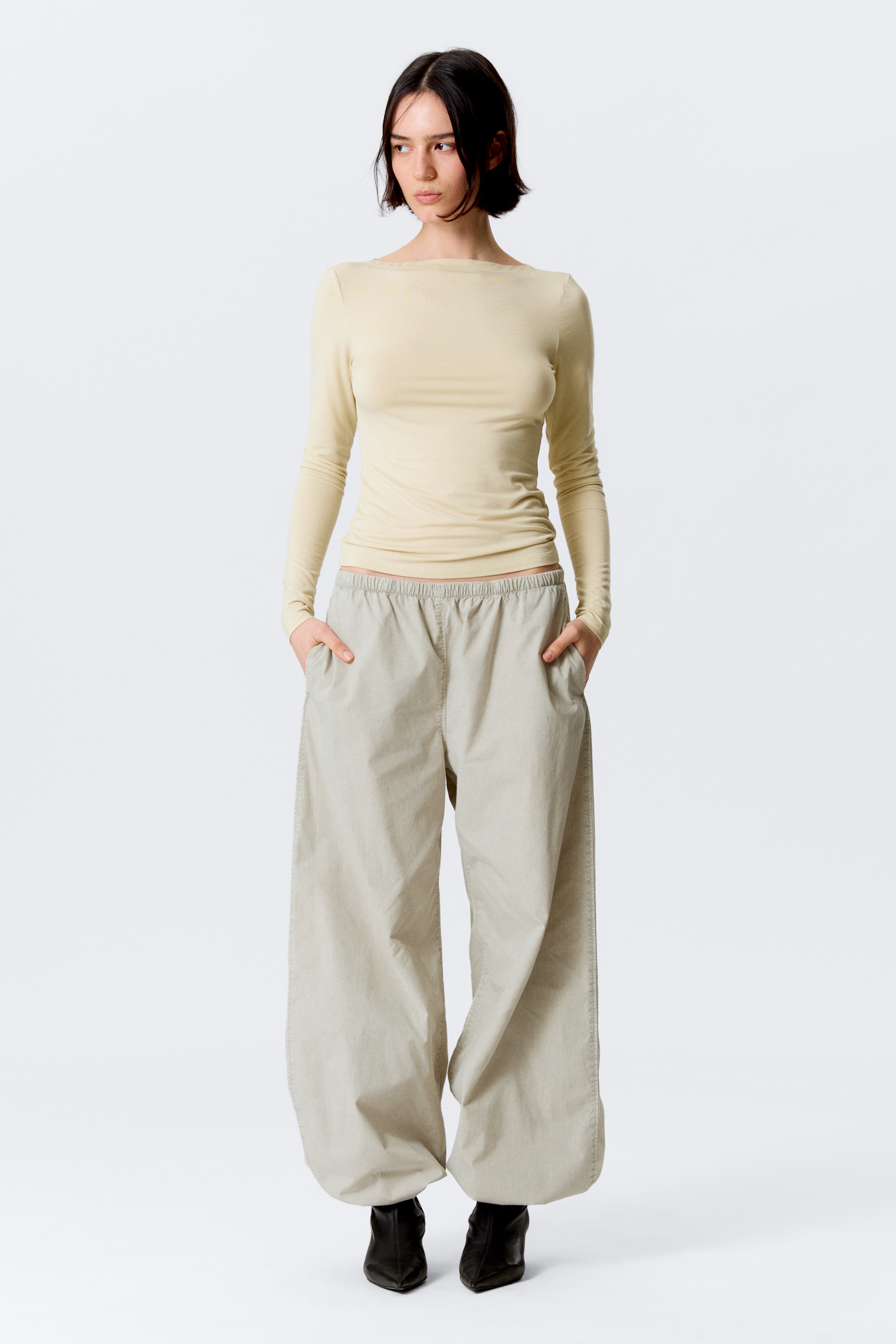 Agrandir l'image: Un haut ajusté de couleur jaune pâle à manches longues avec un col bateau est présenté de face, rentré dans un pantalon ballon beige clair. Le pantalon est conçu avec une taille élastiquée, des poches latérales et des bas de jambes froncés à la cheville, qui tombent sur des bottes noires à bout pointu.