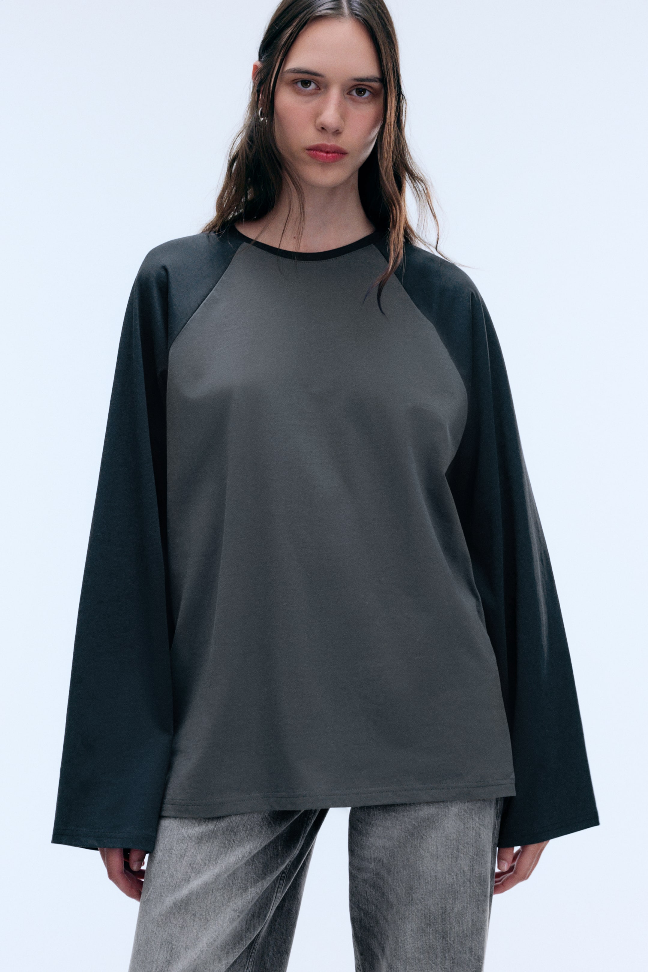 Visualizza l'immagine ingrandita: La modella indossa un top ampio a maniche lunghe con taglio raglan, caratterizzato da un corpo grigio scuro, maniche nere a contrasto e girocollo. Il top presenta maniche dal taglio ampio ed è abbinato a jeans grigi lavati.