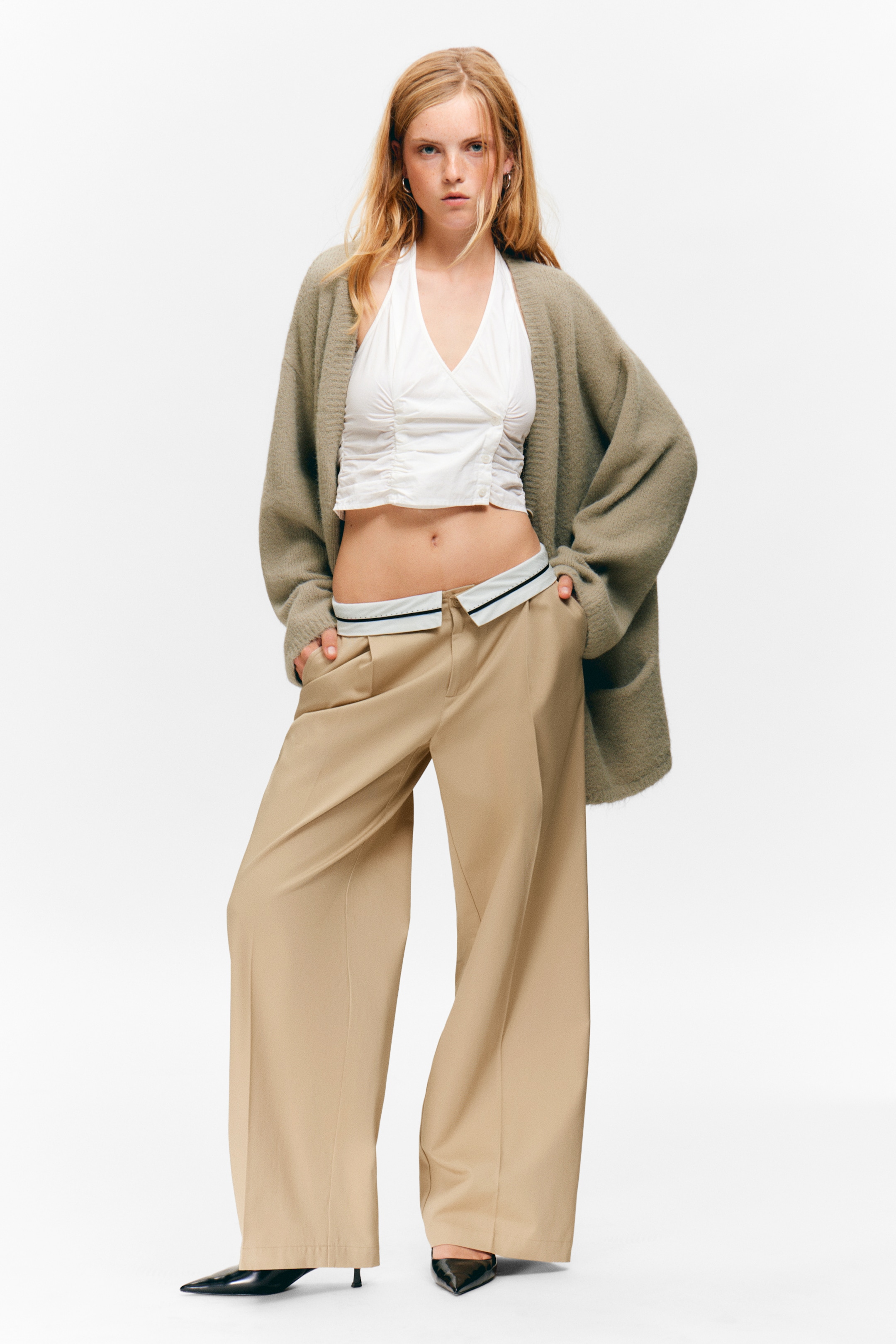 Ingrandisci l'immagine: Un cardigan oversize verde spento aperto sul davanti è drappeggiato su un top bianco corto con scollo all'americana e dettagli arricciati. Pantaloni a gamba larga color cuoio con una cintura interna a contrasto sono indossati, abbinati a décolleté nere a punta con tacco basso.