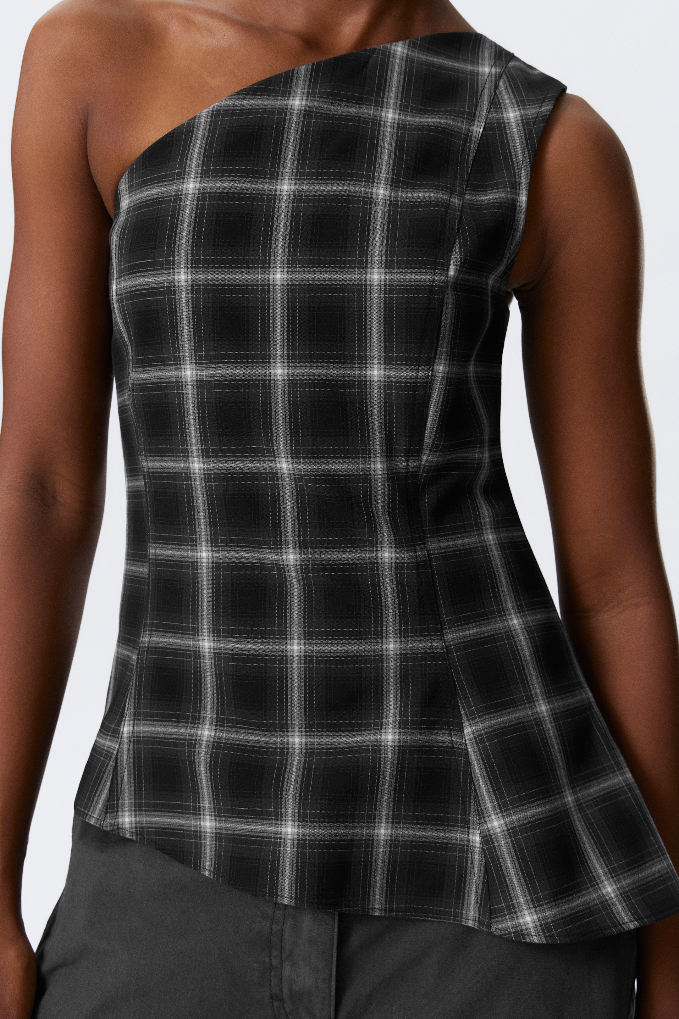 Grösseres Bild ansehen: Ein schwarz-weiß kariertes One-Shoulder-Top wird präsentiert, das ein eng anliegendes Oberteil mit sichtbaren Formnähten aufweist. Das Top ist mit einem asymmetrischen Ausschnitt gestaltet, bei dem eine Schulter bedeckt ist, und einem dezent ausgestellten Schößchen-Saum.