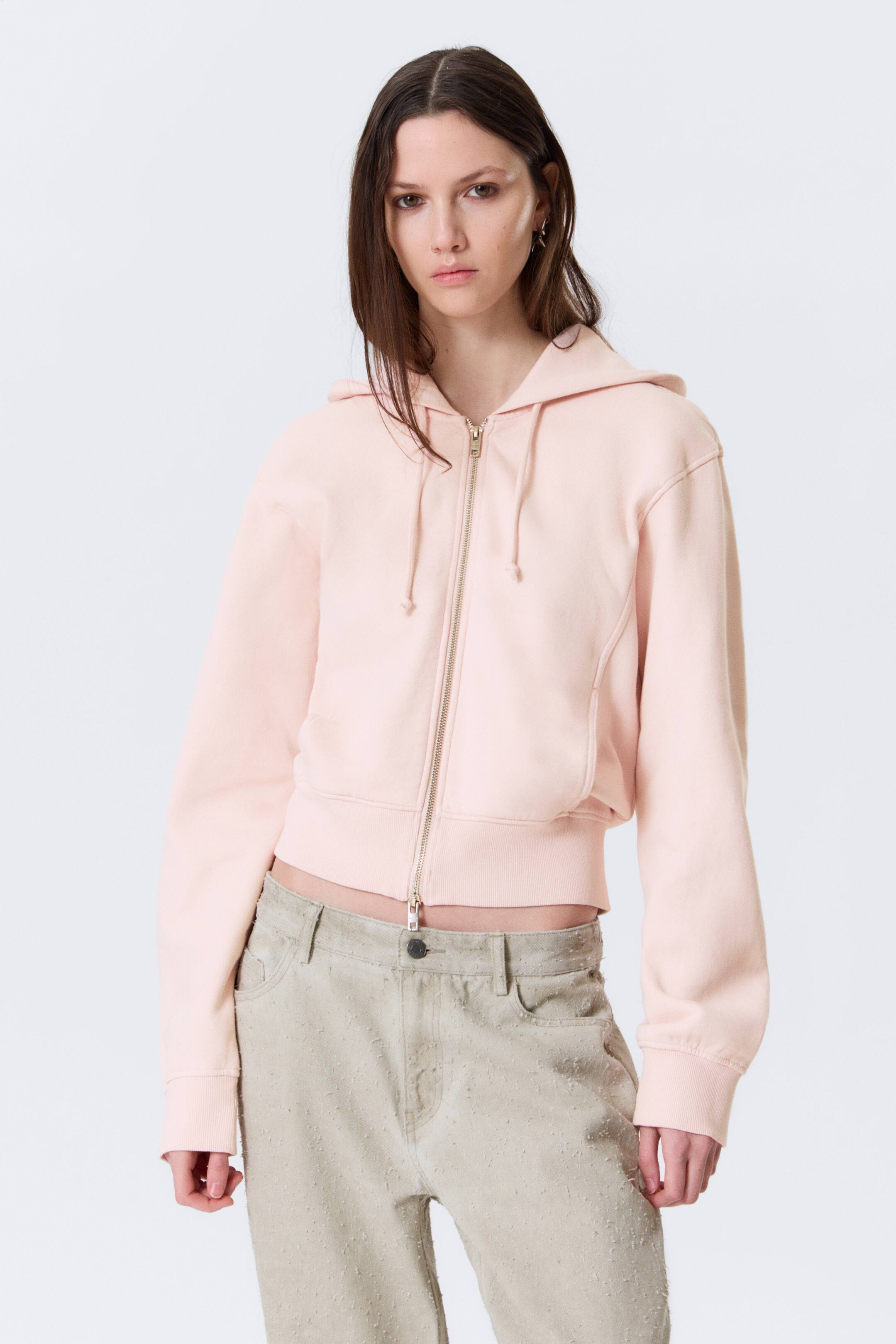 View larger image: A light pink cropped full-zip hoodie is facing forward, featuring a gold-tone zip that is partially unzipped, a hood with drawstrings, and long sleeves with ribbed cuffs. The hoodie also displays a ribbed hem and diagonal seam details on its front.