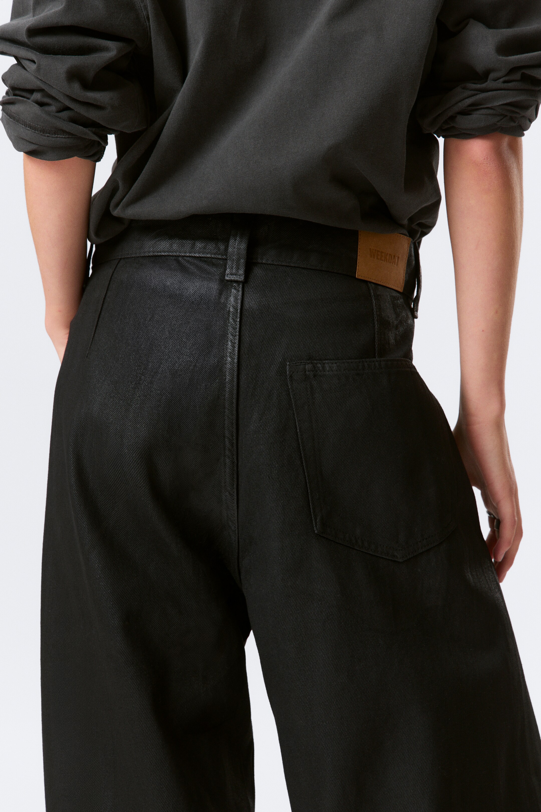 View larger image: Black denim jeans are being presented from the rear, featuring a high waist, belt loops, and a relaxed, wide-leg fit, with a light brown "WEEKDAY" brand patch visible on the right side of the waistband above a patch pocket. A dark long-sleeved top is being worn with the jeans, defining the waistband.