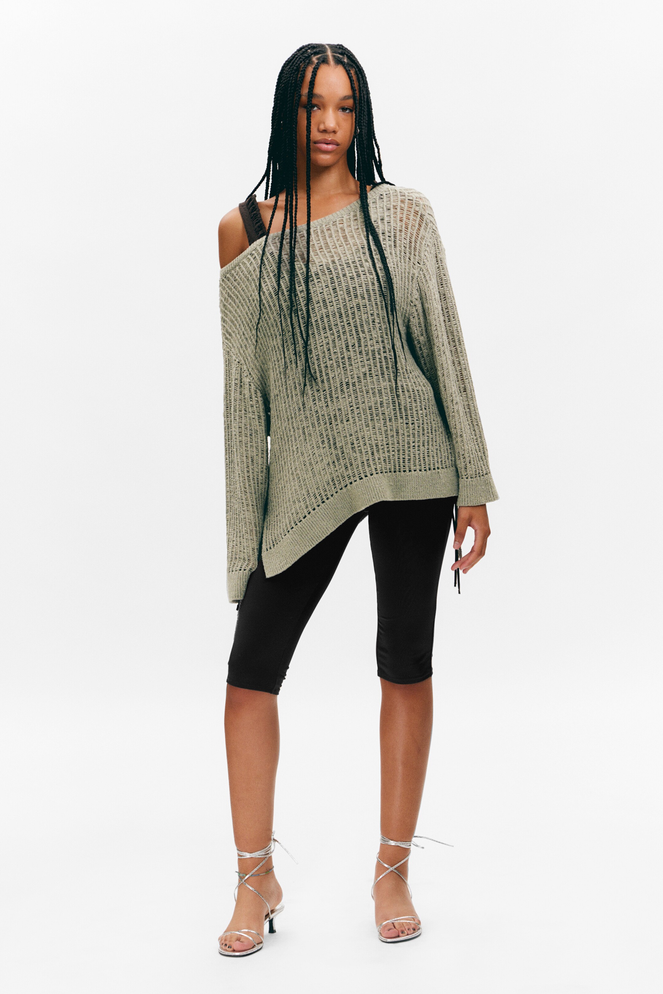 Größeres Bild anzeigen: Ein hellgrüner Open-Knit-Pullover wird aus einer frontalen Perspektive präsentiert, mit einer übergroßen, lockeren Passform und einem asymmetrischen, schulterfreien Ausschnitt auf der linken Seite des Trägers. Seine durchscheinende Konstruktion offenbart einen dunklen Träger darunter, und der Pullover erstreckt sich über schwarze Shorts mit einem asymmetrischen Saum.