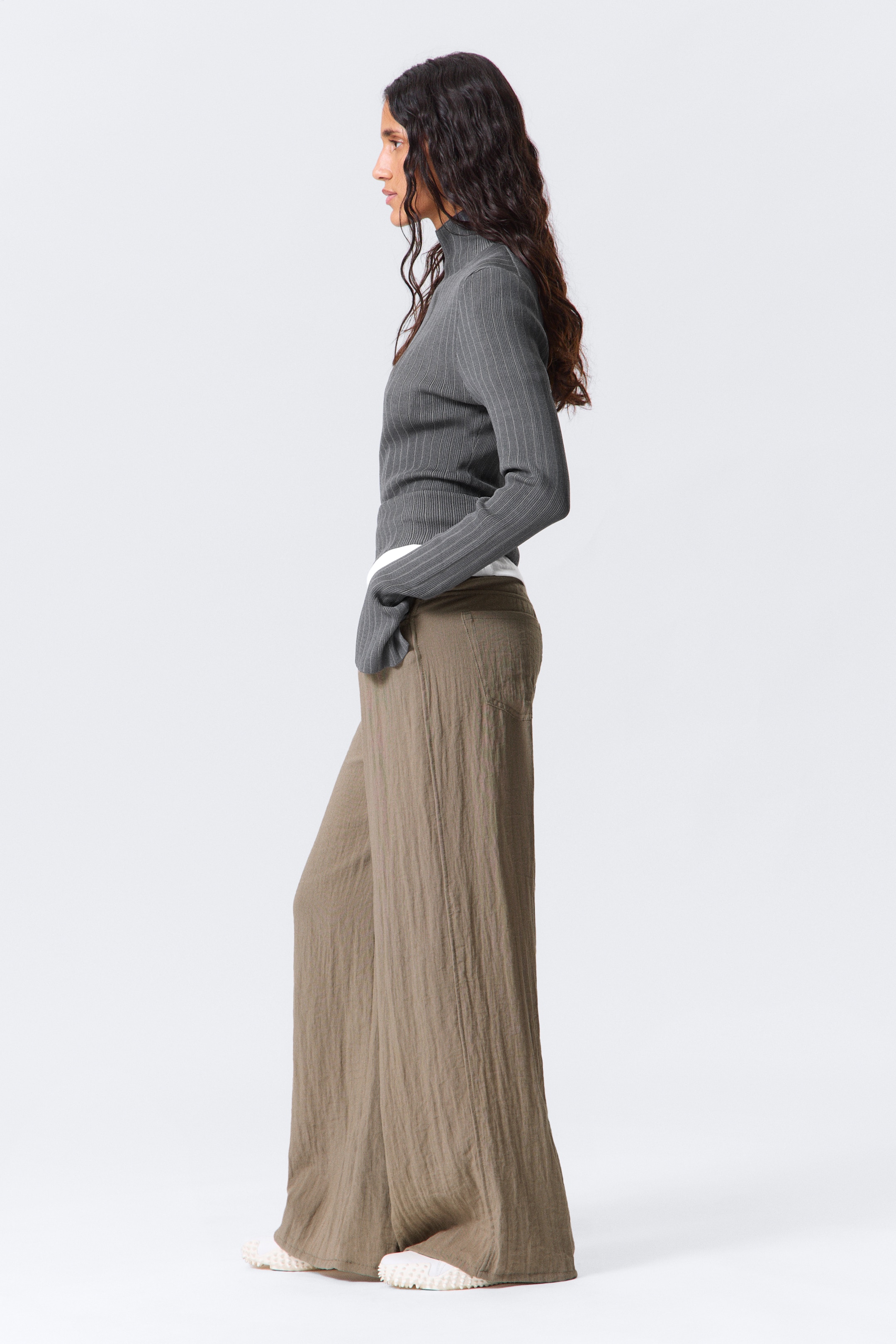 View larger image: A grey, ribbed, high-neck top is being shown in a side profile, featuring a slim fit and fine vertical ribbing, layered over a white garment. A pair of wide-leg khaki pants with a textured fabric are being worn below, showcasing their loose fit and full length, resting over white athletic shoes.