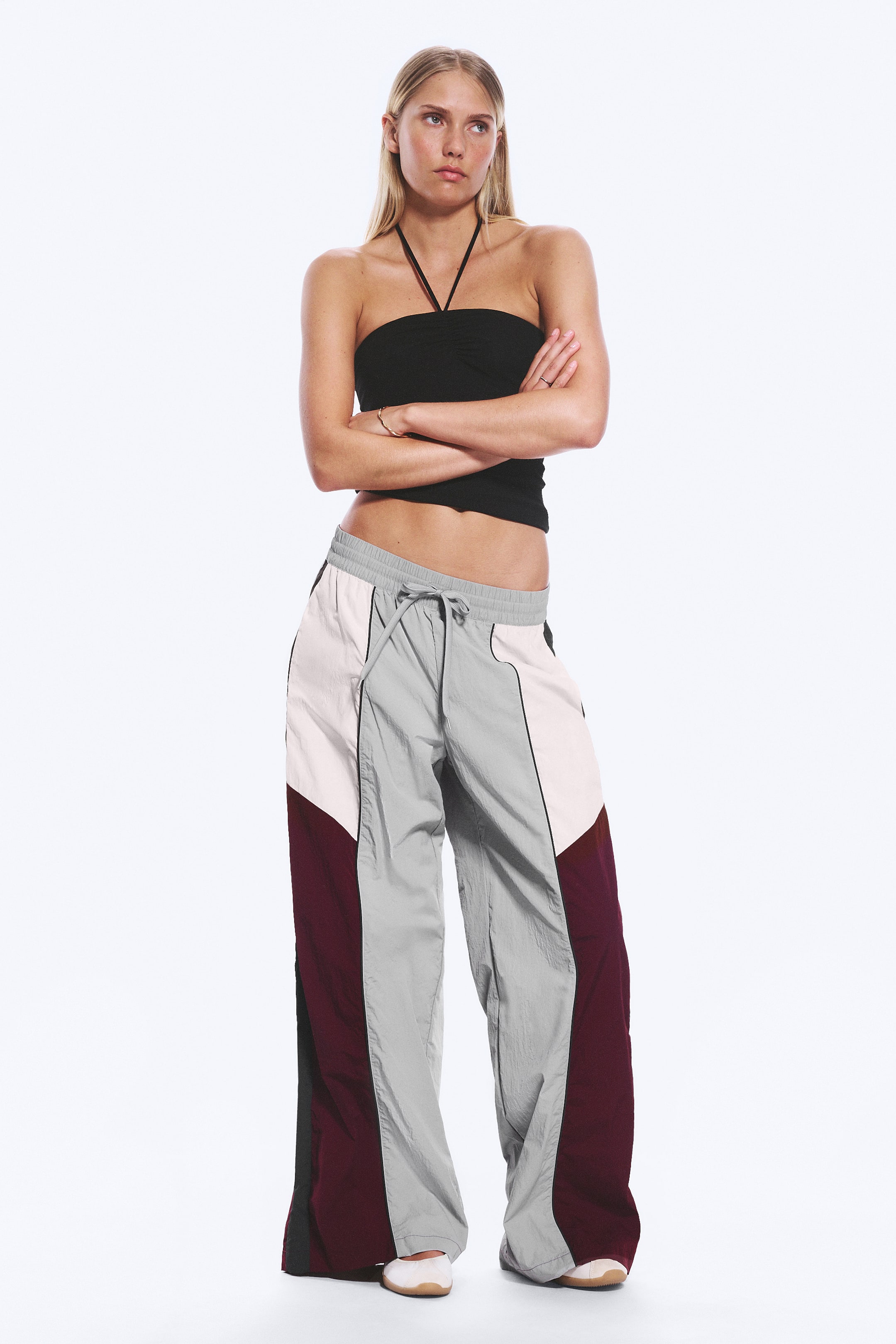 Se større billede: En sort bandeau-top med en halterneck-strop er stylet med bredbenede trackbukser. Bukserne har farveblokpaneler i lysegrå, off-white og bordeaux, fremhævet med sorte lodrette pipings og en elastisk snøre i taljen.