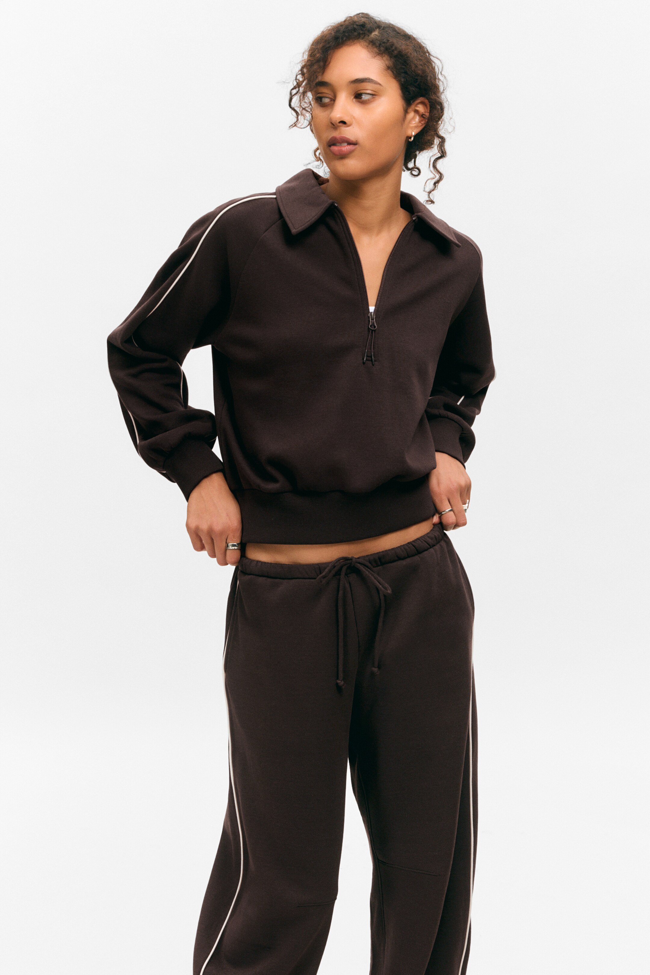 Ingrandisci l'immagine: Un set da salotto marrone scuro viene presentato frontalmente, caratterizzato da una felpa a collo alto con una chiusura a zip da un quarto, piping bianco che accentua le maniche raglan, e polsini e orlo a coste. Anche i pantaloni della tuta marrone scuro abbinati vengono mostrati, caratterizzati da una vita con coulisse e piping bianco che dettaglia le cuciture laterali.
