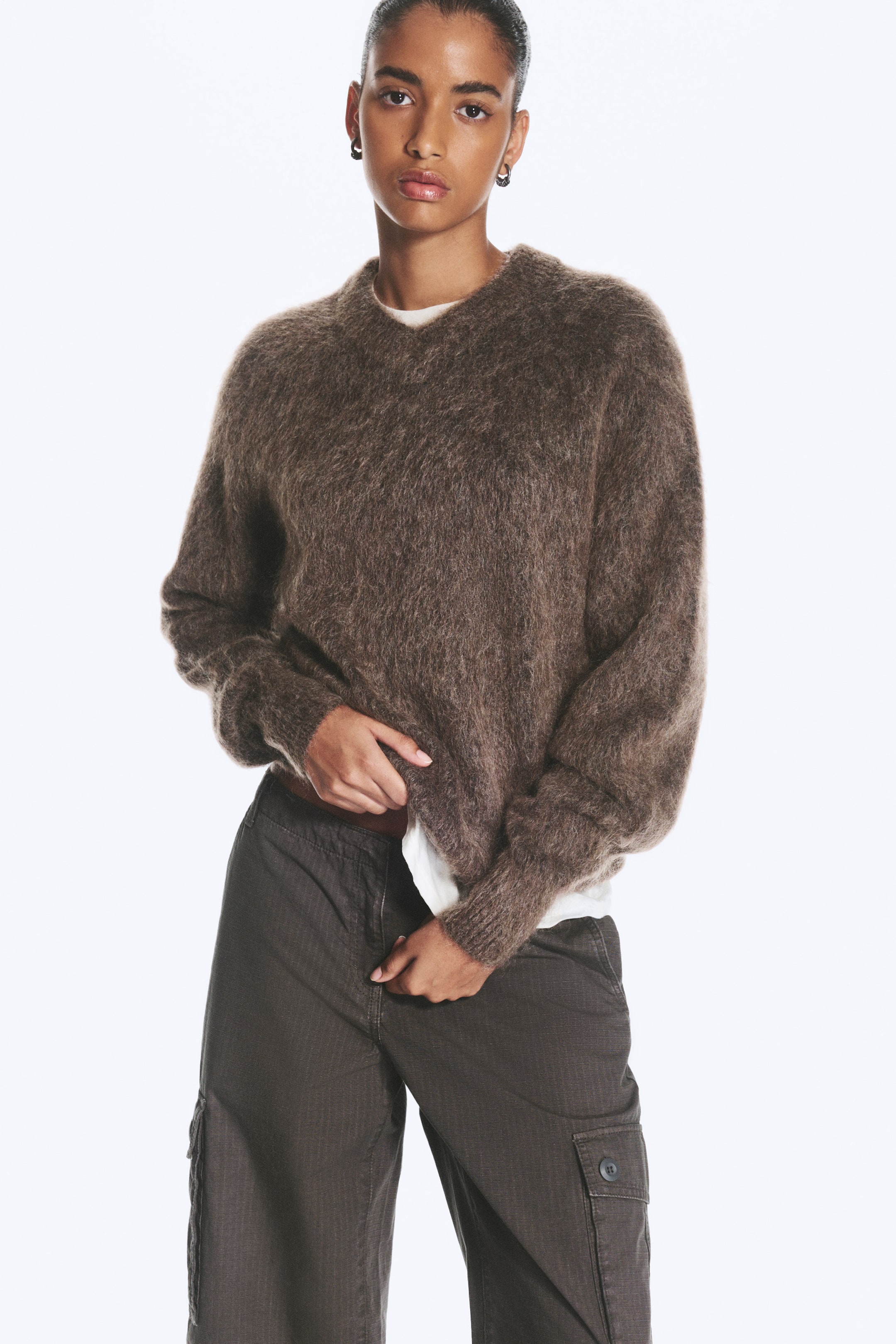 Grösseres Bild ansehen: Ein brauner, gestrickter Pullover aus Mohairmischung wird vom Model getragen, das den Saum mit den Händen anhebt, um darunter ein weisses T-Shirt zu zeigen. Der strukturierte Pullover mit langen Ärmeln verfügt über Rippbündchen und ist mit dunklen Cargo-Hosen kombiniert.