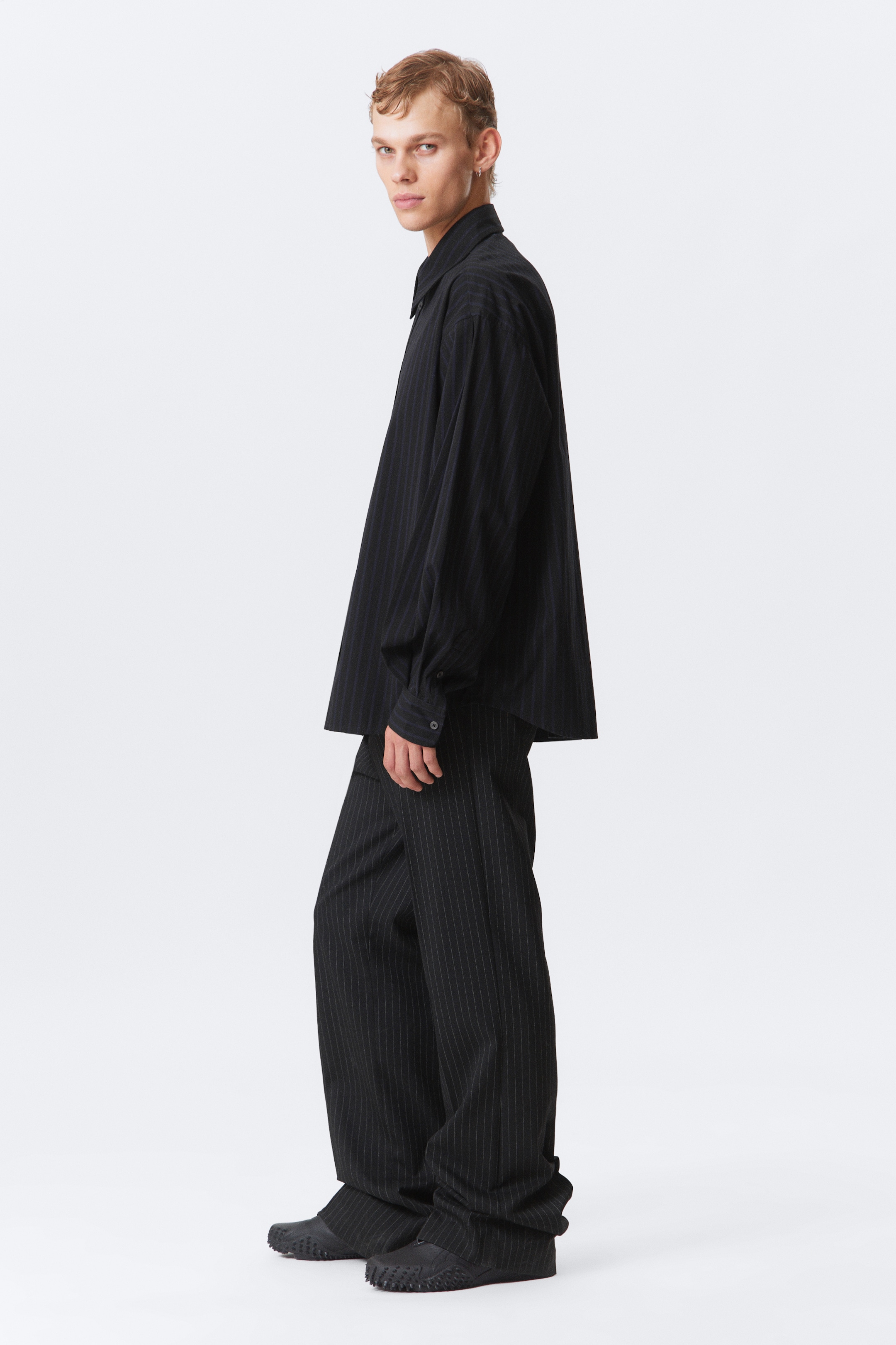 Ver imagen más grande: Una camisa de rayas negras de talla grande y pantalones anchos a juego se presentan de perfil lateral. La camisa presenta un cuello clásico y puños abotonados, mientras que los pantalones se agrupan en los tobillos sobre zapatos negros con detalles tachonados en la punta.