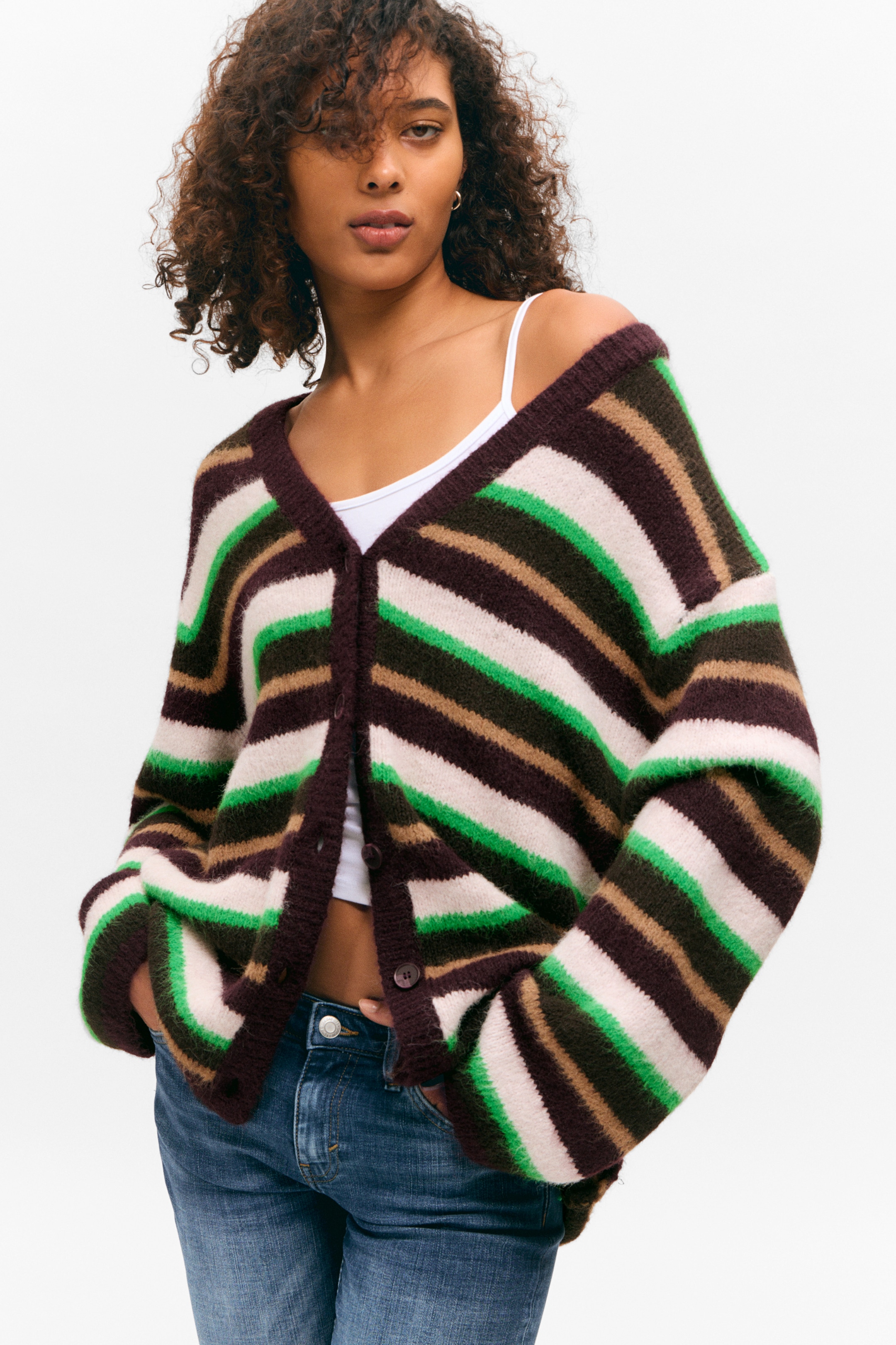 Größeres Bild anzeigen: Ein V-Ausschnitt-Cardigan, gestreift in Dunkelbraun, Hellbraun, Grün und Off-White, wird von dem Model getragen. Der übergroße Cardigan zeichnet sich durch fallende Schultern, eine Knopfleiste mit sichtbaren Knöpfen, gerippte Trimmungen und Vordertaschen aus, in die das Model ihre Hände steckt, über einem weißen Camisole und blauen Jeans geschichtet.