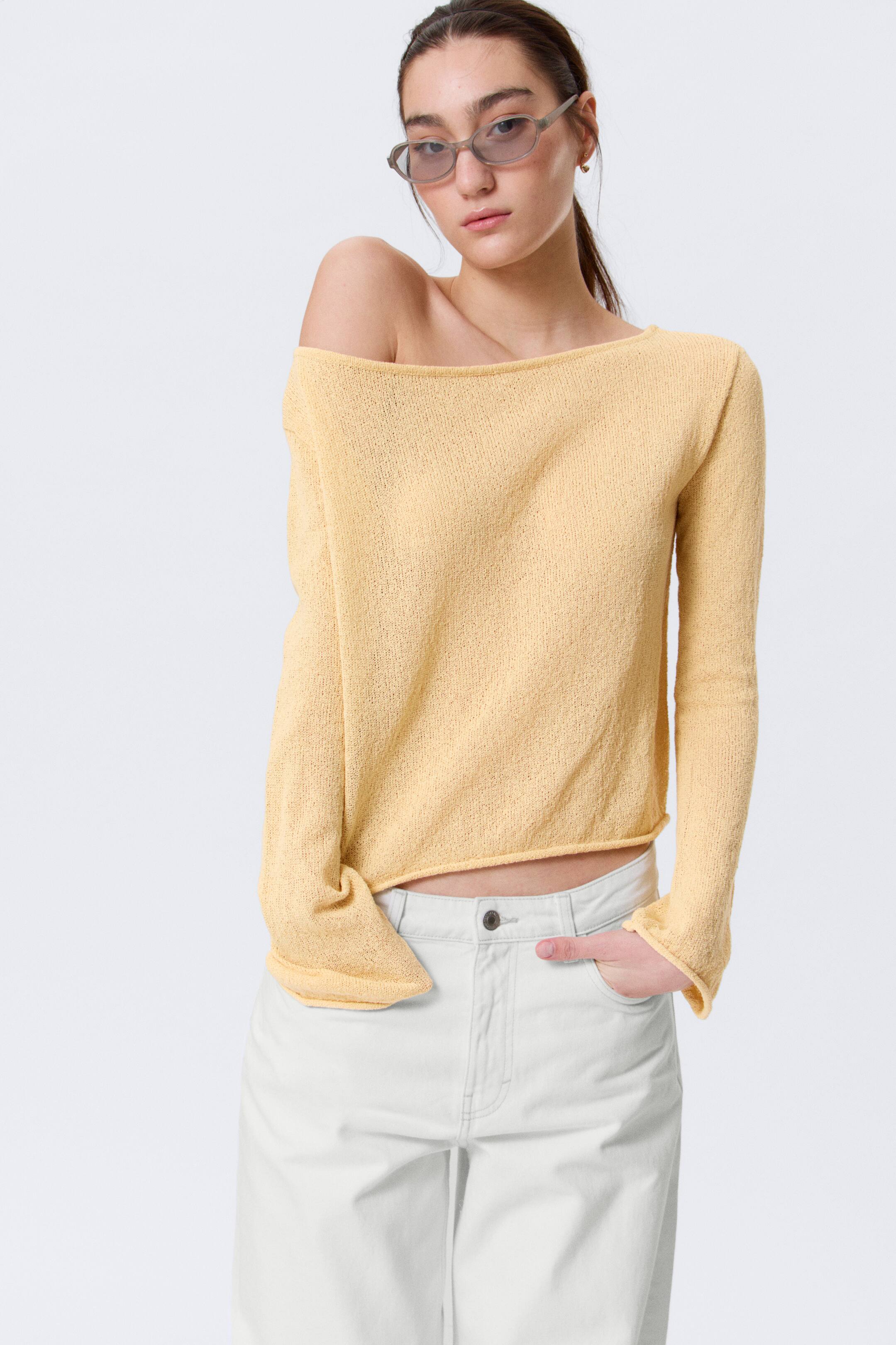 Ingrandisci l'immagine: Un top a maglia color giallo chiaro con scollo a barchetta, caratterizzato da una lavorazione a maglia testurizzata e maniche lunghe, indossato su una spalla e parzialmente infilato in pantaloni a gamba larga color crema chiaro.
