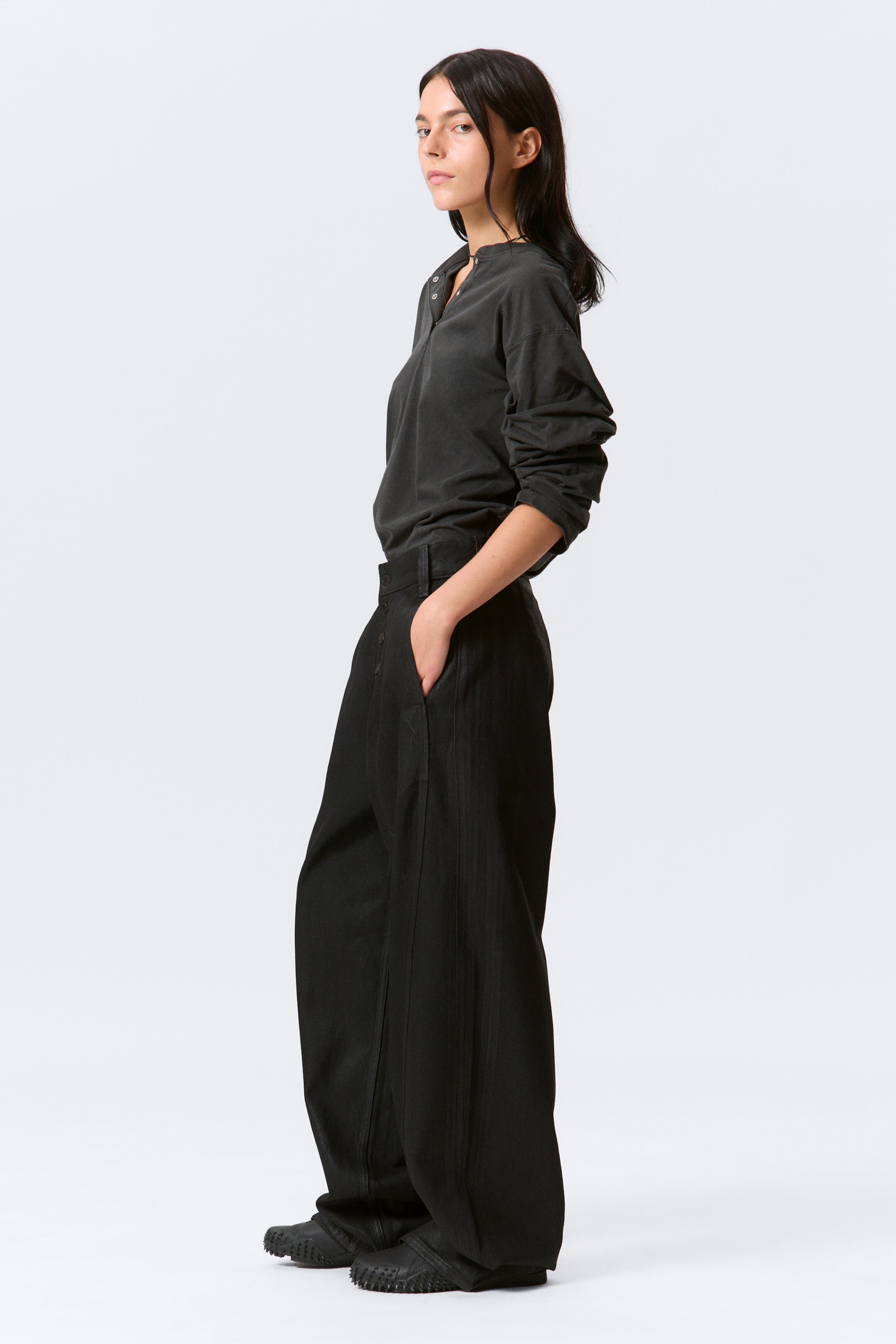 View larger image: A dark grey long-sleeved Henley shirt, featuring a partial button placket and relaxed sleeves, is being worn tucked into black wide-leg pants. The pants are showcasing prominent vertical seam details down the front of each leg and are presenting a high-waisted, flowing silhouette from a side-facing angle.
