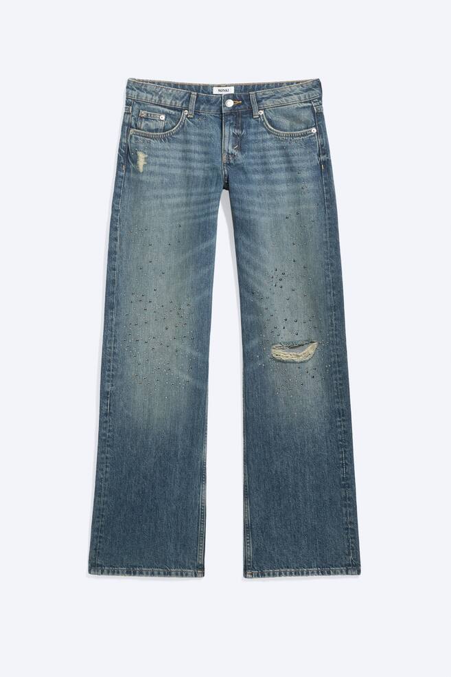 Unos vaqueros de denim azul de lavado medio se muestran de frente, con una silueta de pierna recta y adornos de tachuelas plateadas dispersos por la parte delantera de ambas perneras. Los vaqueros presentan detalles desgastados, incluido un pequeÃ±o parche deshilachado cerca del bolsillo derecho y un desgarrÃ³n horizontal en la rodilla izquierda. - 1